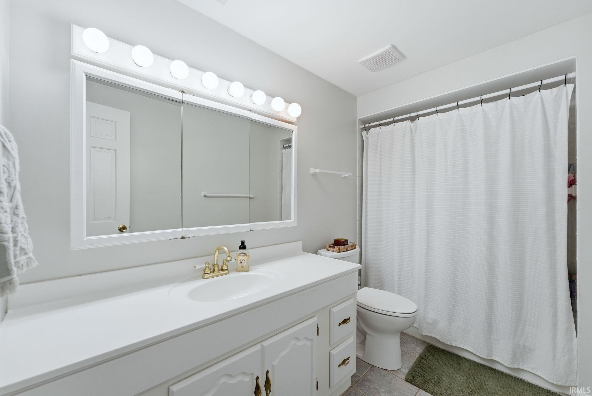Bathroom featuring a white vanity with integrated sink, brass-finish faucet, and a large mirror with Hollywood-style lighting