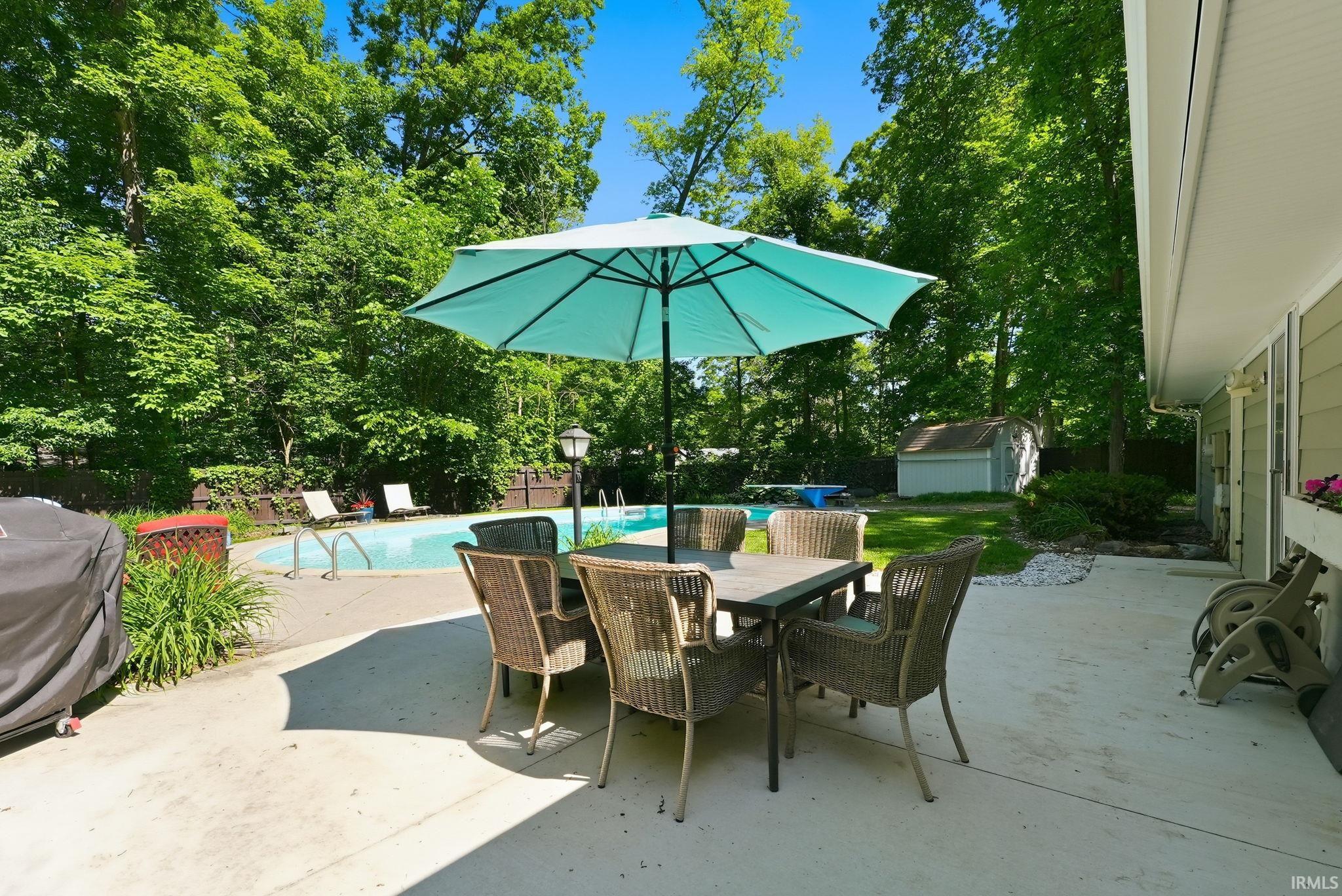 Concrete patio with a dining area, featuring a fenced-in swimming pool, lush mature trees, and a detached storage shed
