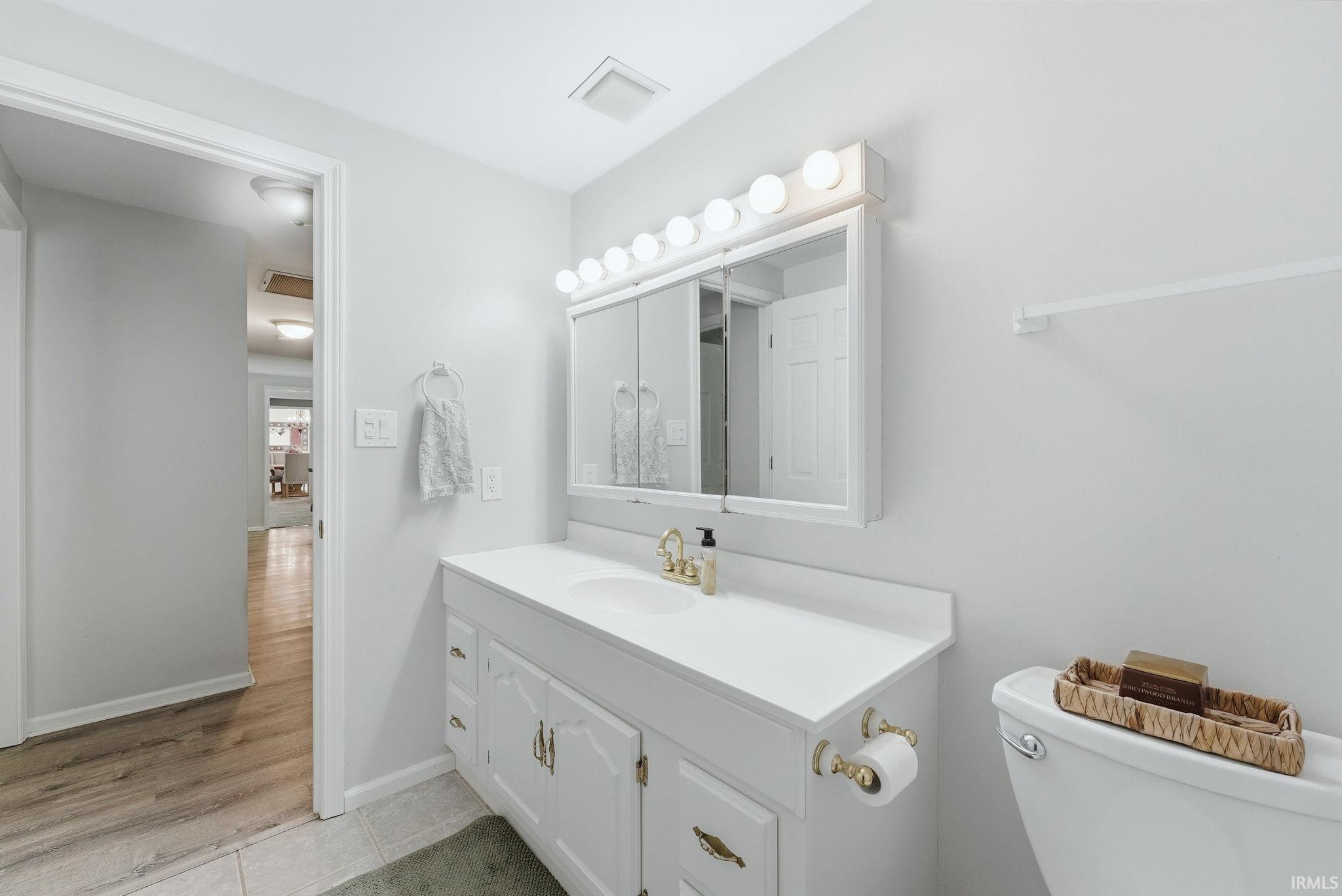 Bathroom with a white vanity featuring gold-toned hardware, a white countertop with an integrated sink, and a wall-mounted medicine cabinet with integrated lighting