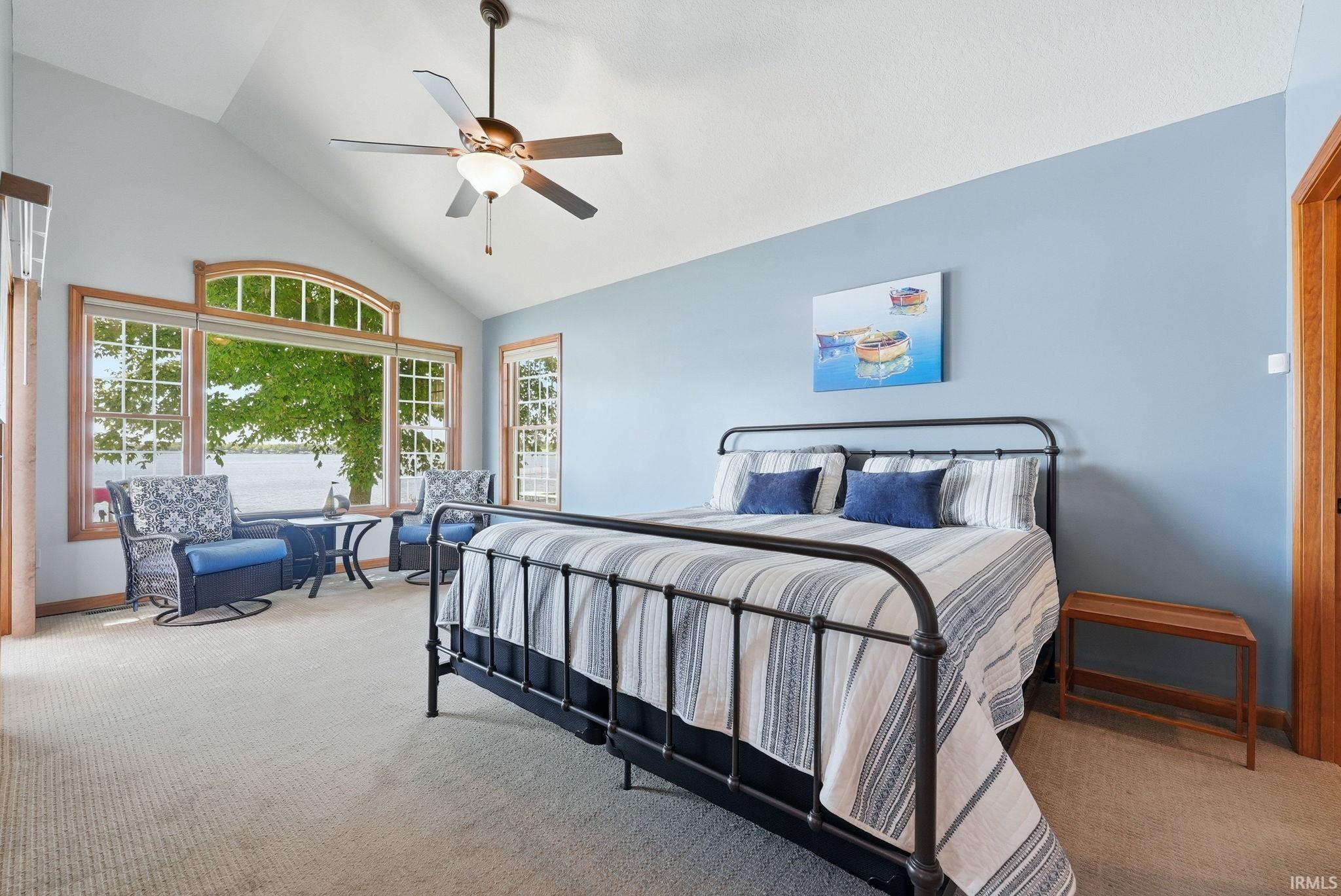 Vaulted ceiling room featuring a large arched window with water scenery, a ceiling fan, light-colored carpeting, and natural wood trim throughout