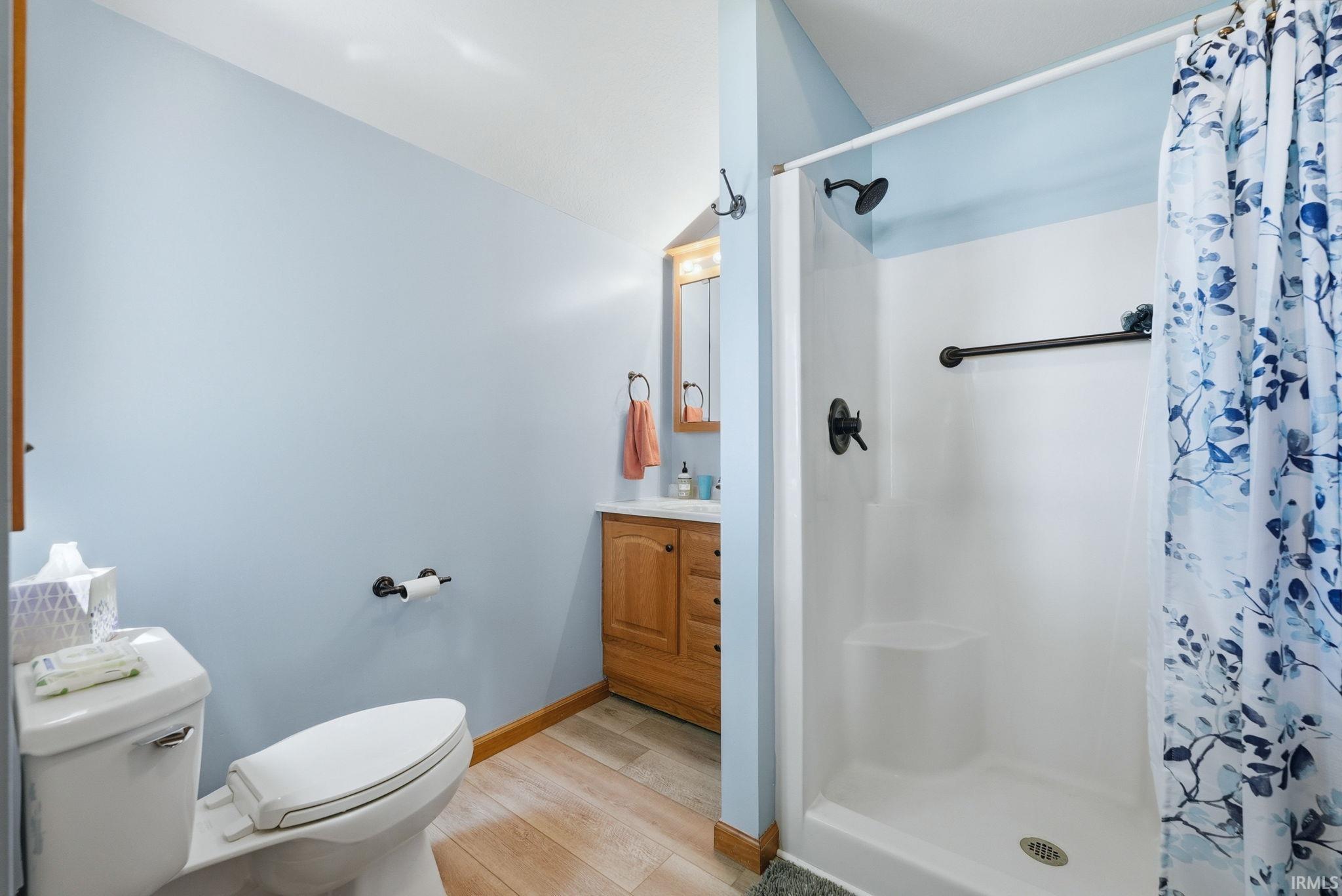 Three-piece bathroom featuring a stand-up shower with matte black fixtures, a single vanity with wood cabinetry, and wood-finish flooring