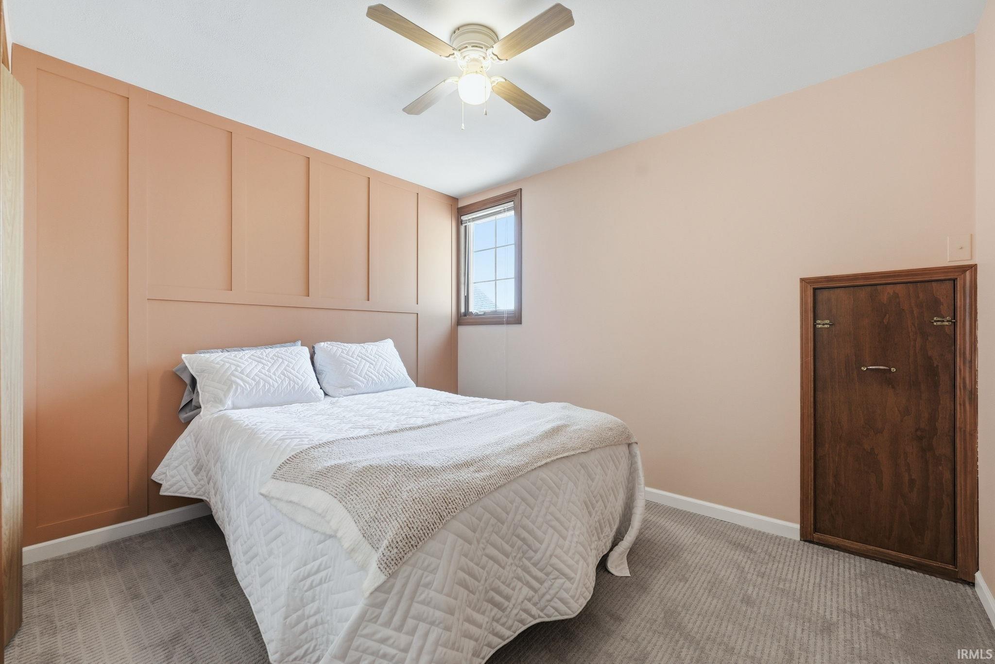 Bedroom featuring an accent wall with vertical paneling, light gray carpeting, a single window with wood trim, a ceiling fan with integrated lighting, and a built-in wood cabinet