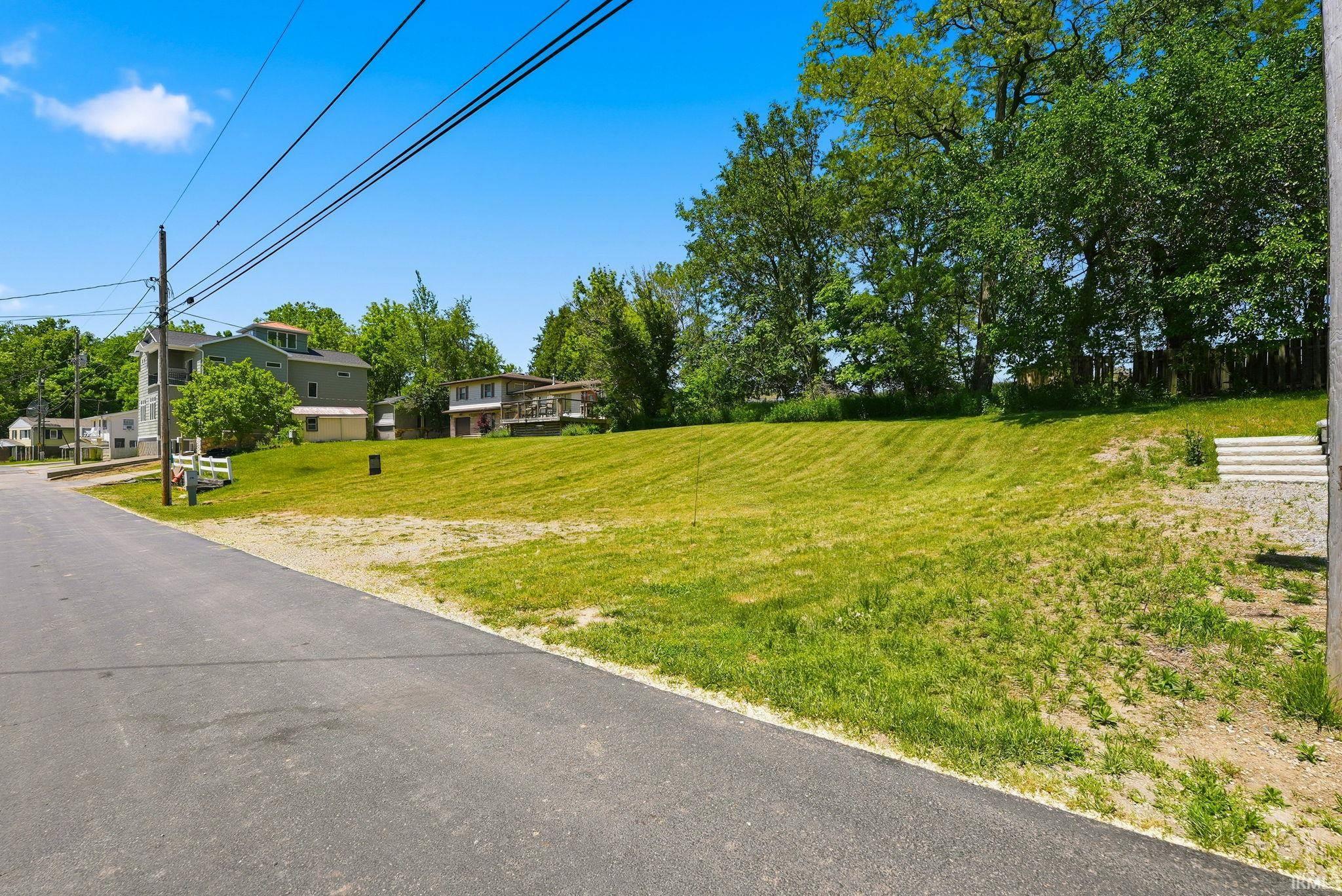 Expansive grass lot featuring a gently sloping terrain, asphalt road frontage, and established trees in the background
