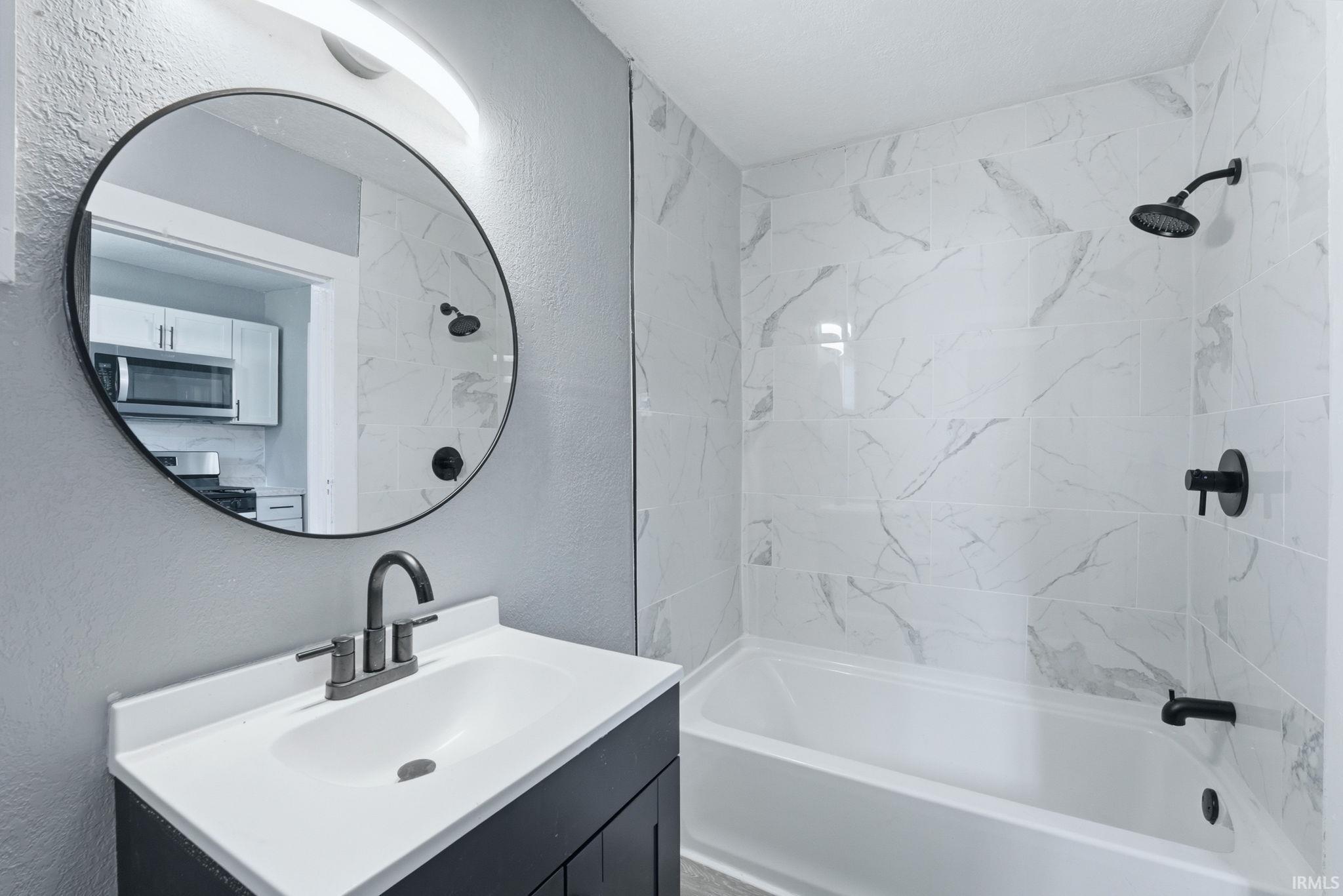 Bathroom featuring a vanity with a white countertop and dark cabinetry, a round mirror, and a tub with white and gray tiled surround
