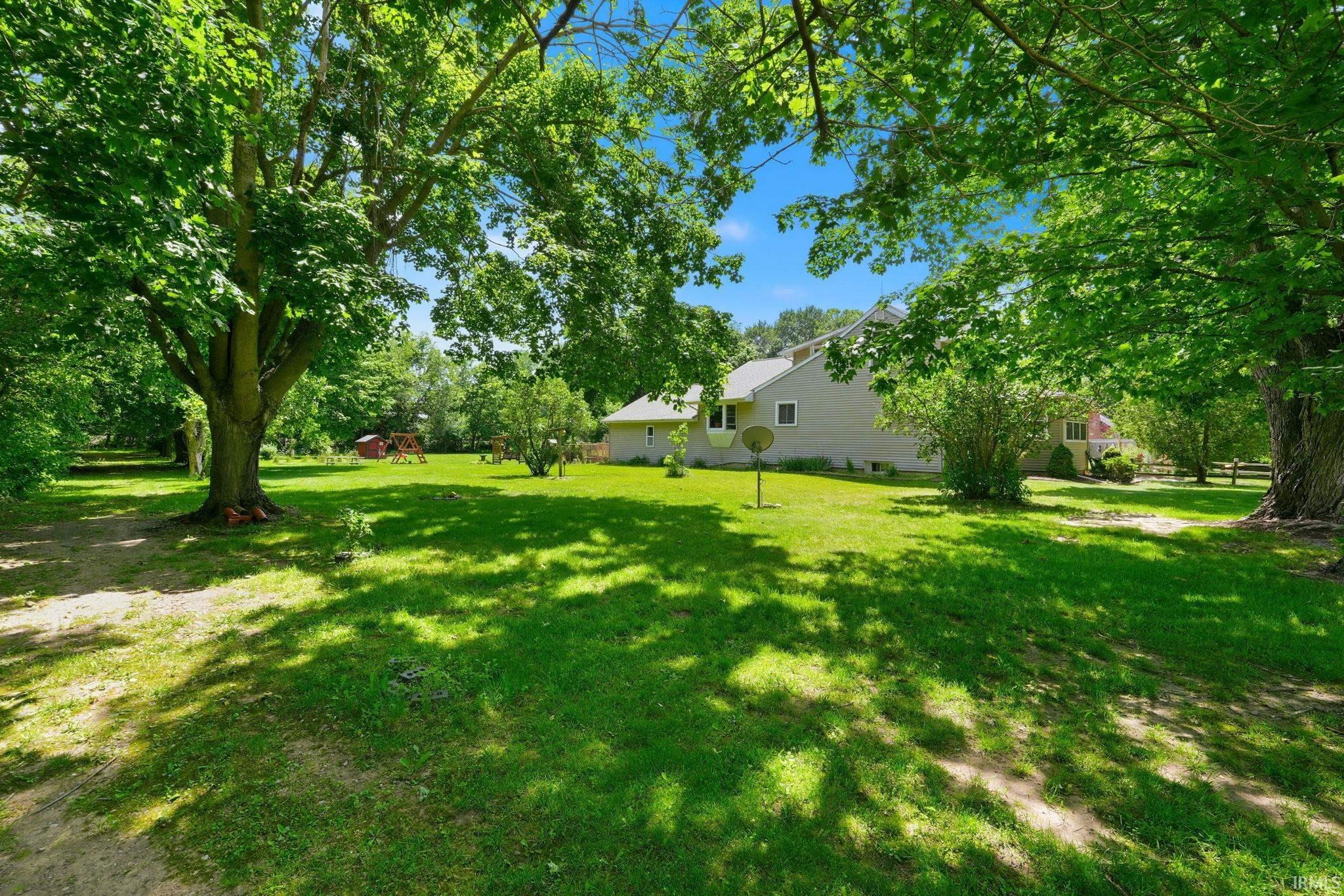 Expansive green lawn framed by mature trees, featuring a light-colored siding residence with multiple windows and a visible fence line