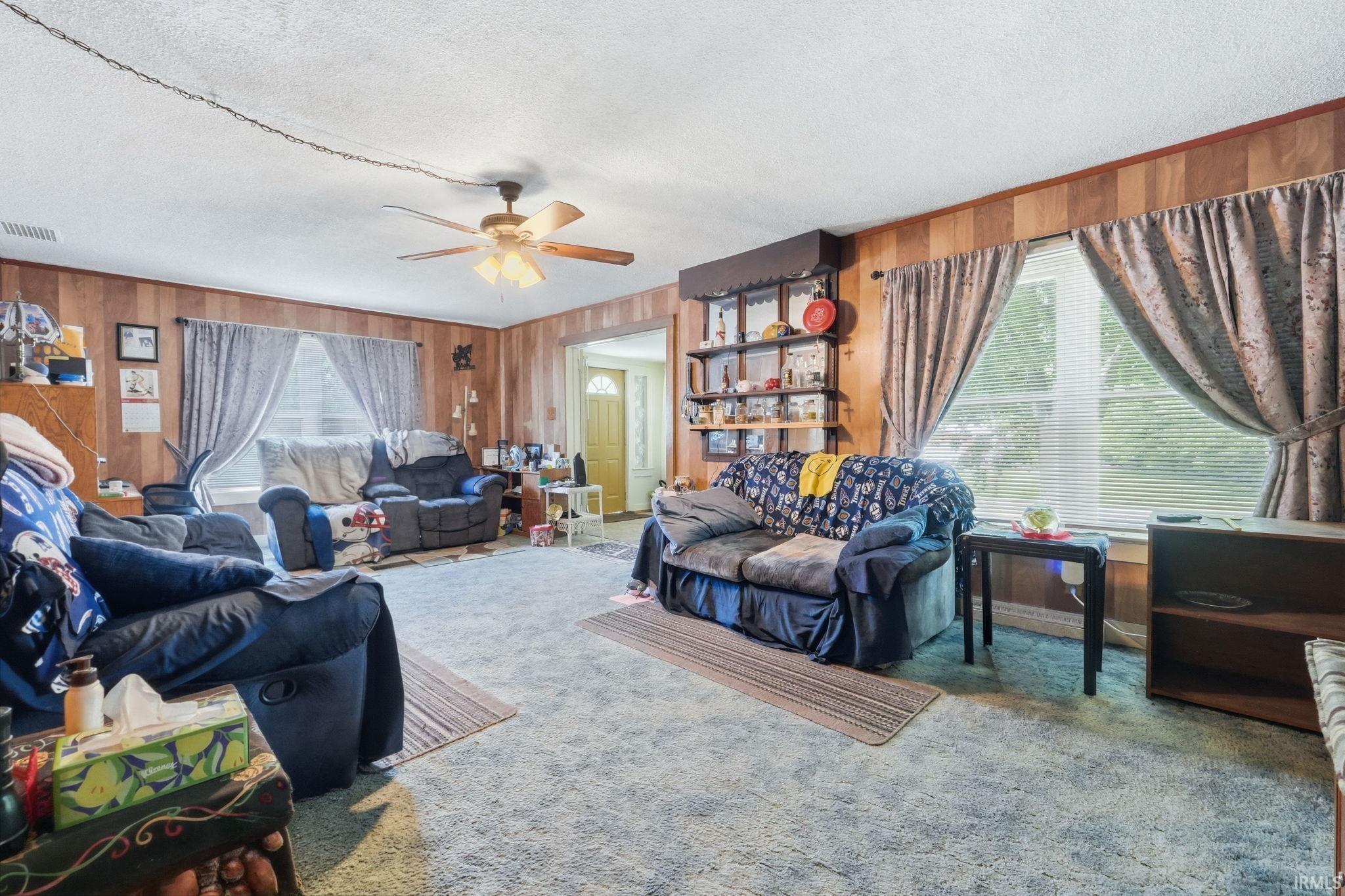 Spacious living area featuring wood-paneled walls, a ceiling fan, and carpeted flooring