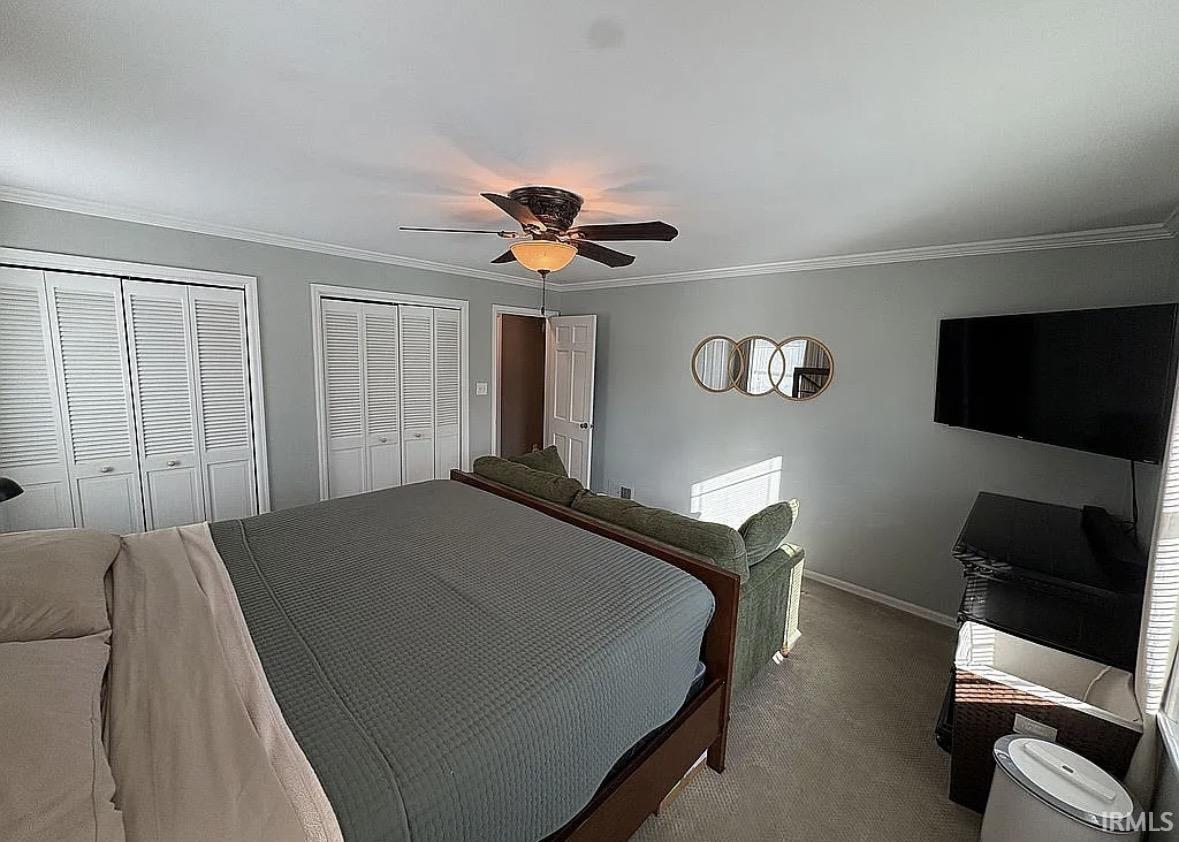 Spacious sleeping area featuring two sets of bi-fold louvered closet doors, crown molding, a ceiling fan with integrated lighting, and neutral carpeting