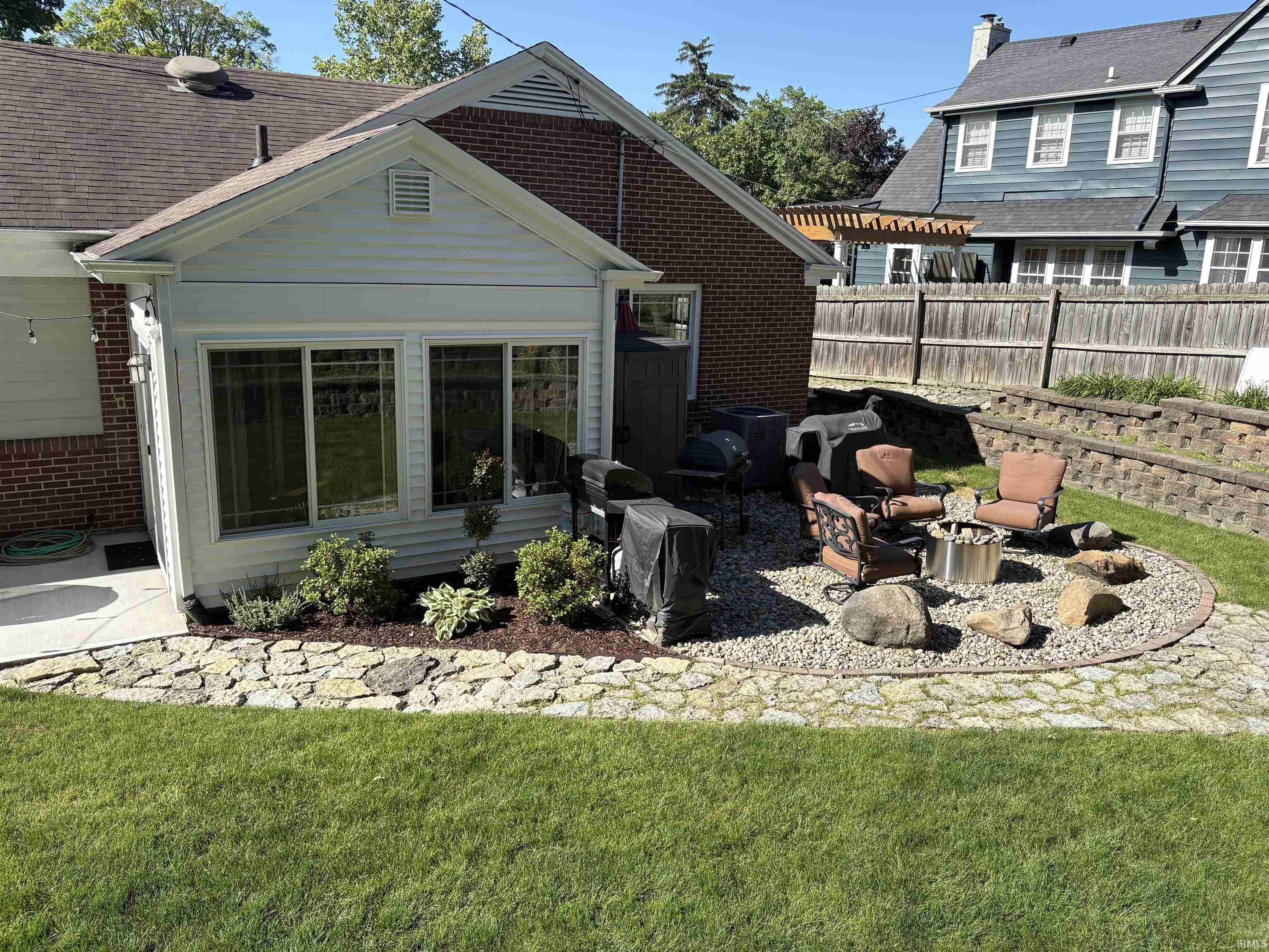 Expansive outdoor living area featuring a sunroom addition with multiple windows, a flagstone and gravel patio with a built-in fire pit, tiered retaining walls, and a manicured lawn