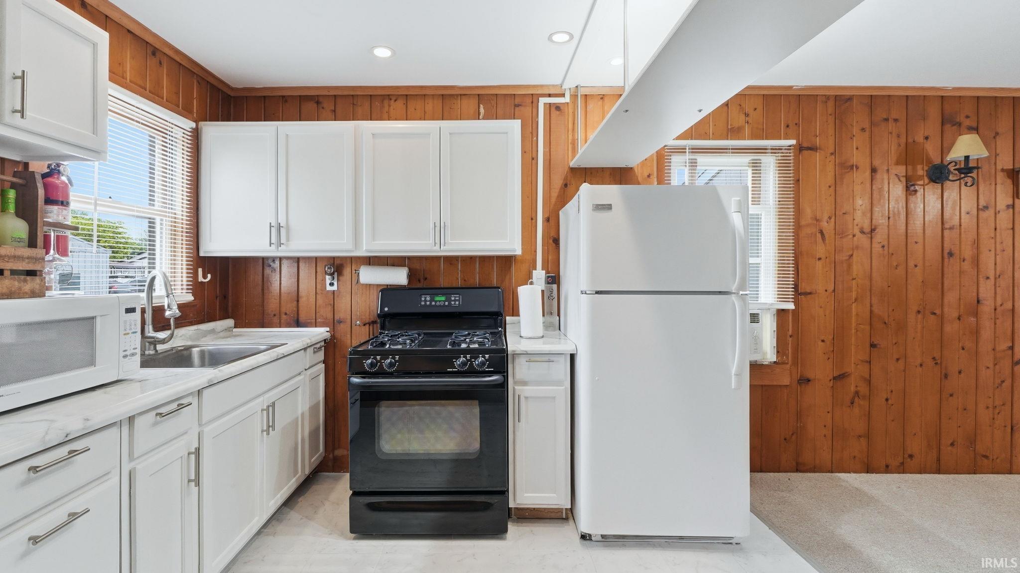 Kitchen featuring wood-paneled walls, white cabinetry with silver-tone hardware, a stainless steel sink, black range, and light-toned flooring