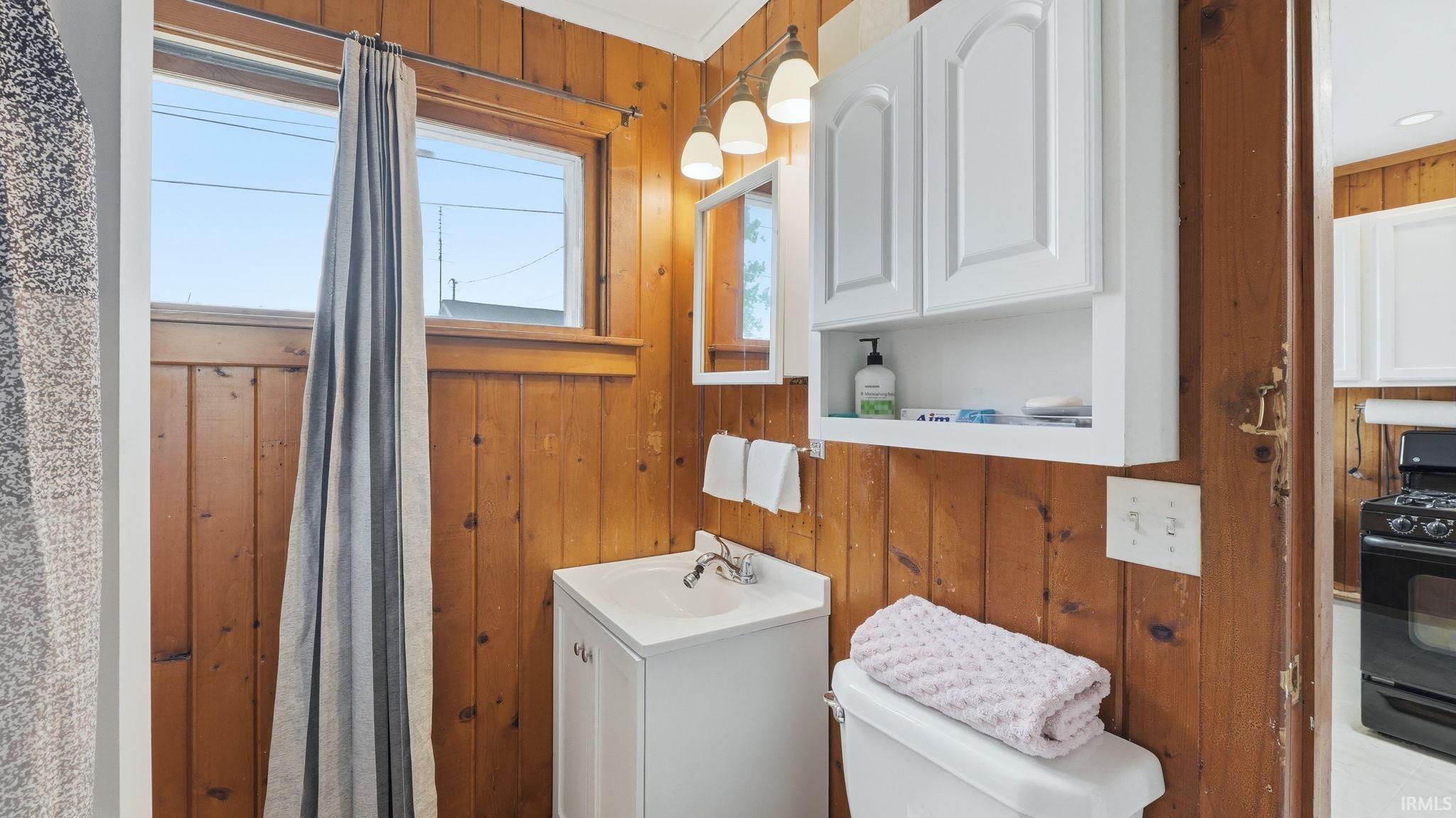 Bathroom featuring wood paneling, a single window, a white vanity with an integrated sink, a mirrored medicine cabinet, and an overhead lighting fixture