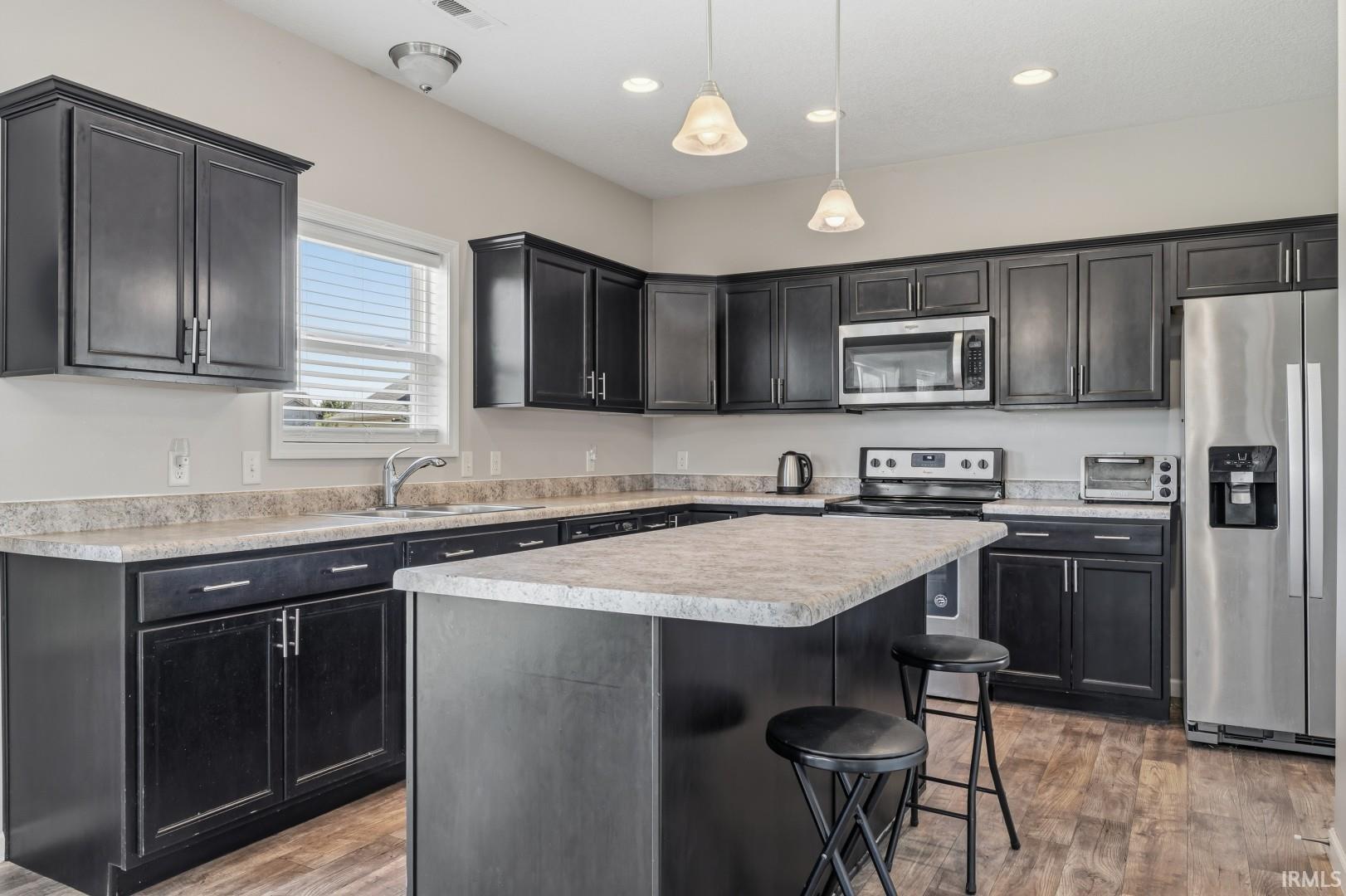 Kitchen featuring dark wood-finish cabinetry, light-toned countertops, a central island with bar seating, stainless steel appliances, and wood-finish flooring