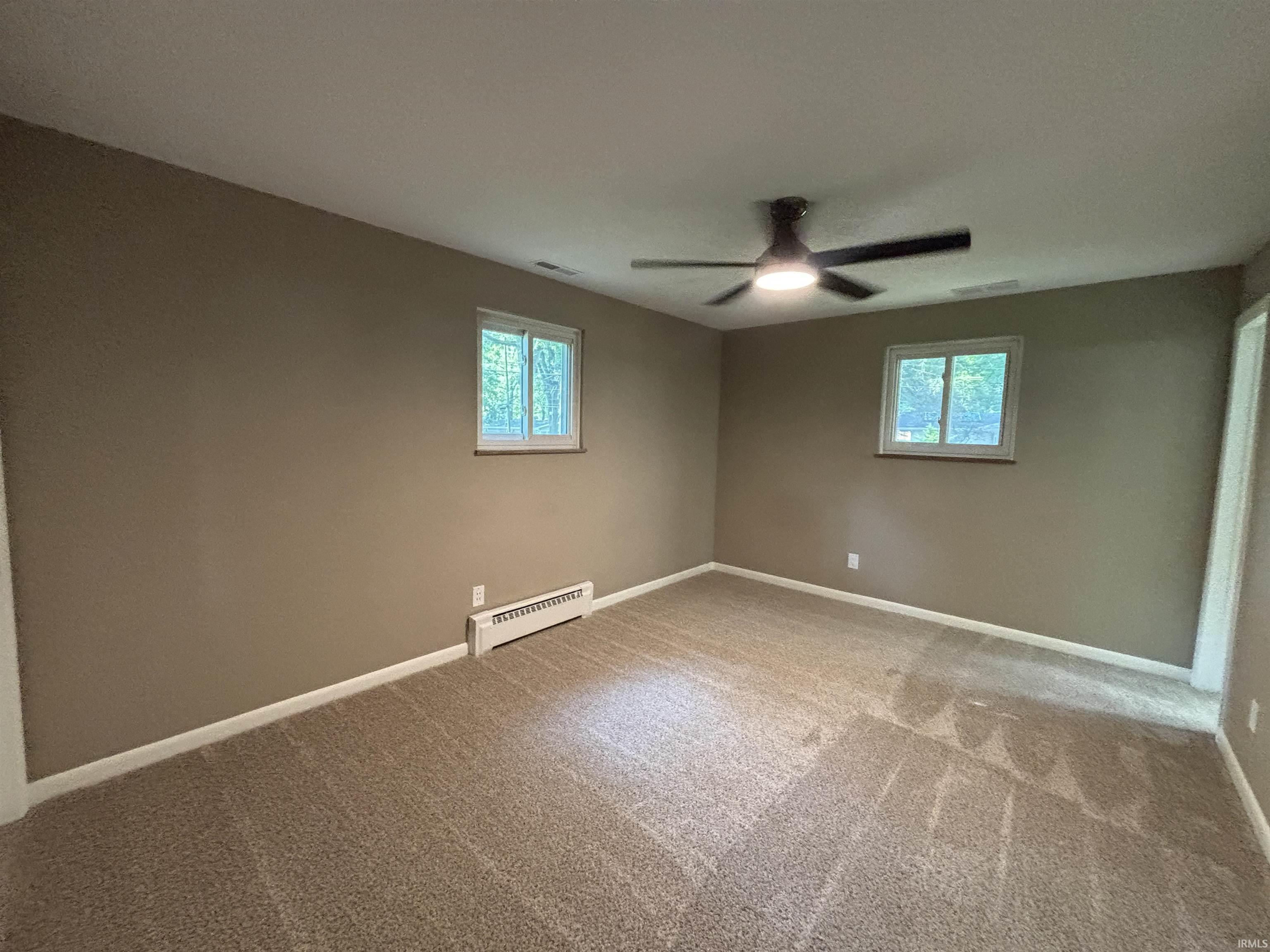 Neutral-toned room featuring a textured carpet, two windows with white trim, a modern ceiling fan with integrated lighting, and white baseboards