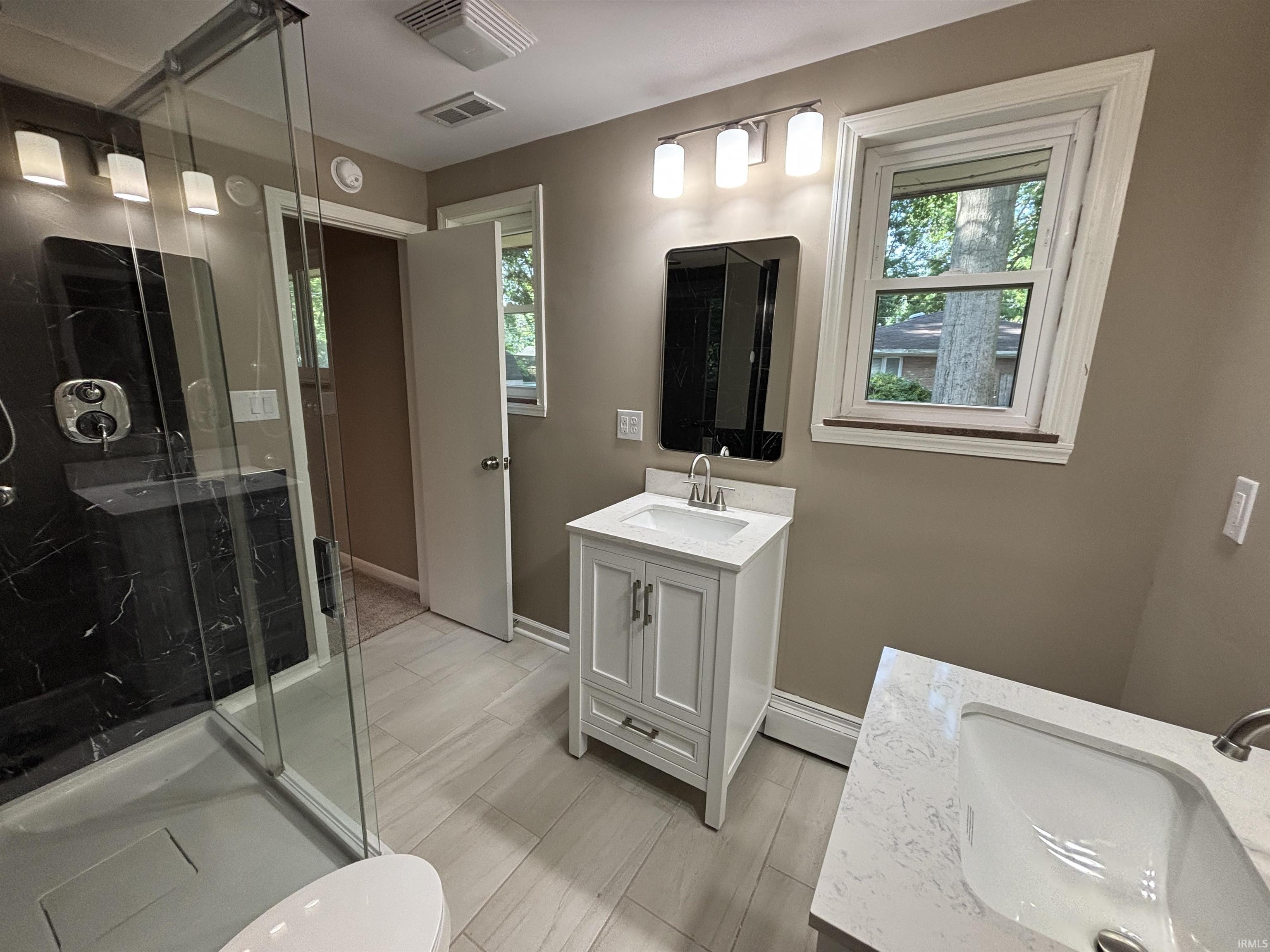 Contemporary bathroom featuring a glass-enclosed shower with black marble-look walls, two white vanity cabinets with integrated sinks, and light wood-finish flooring
