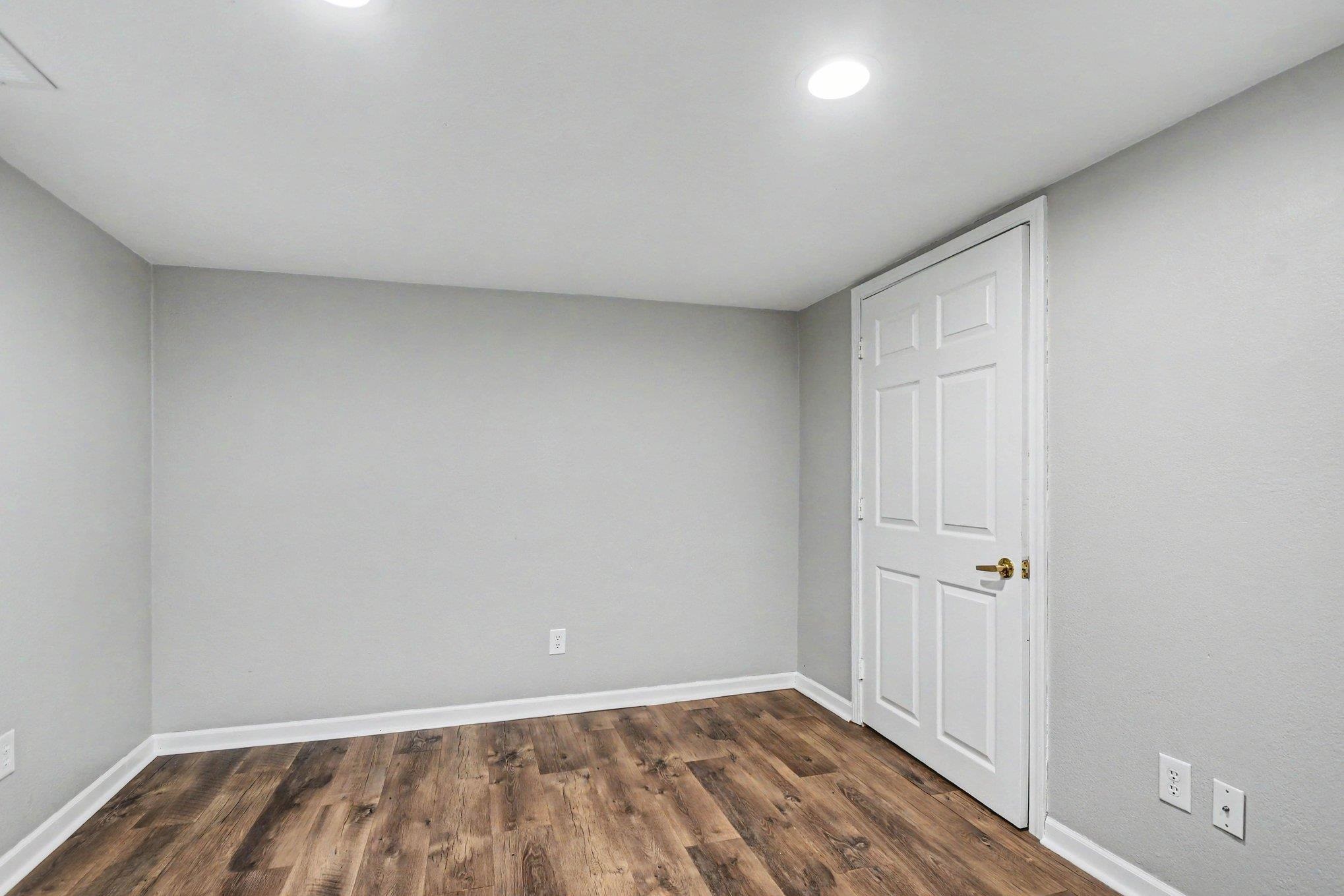 Room featuring wood-finish flooring, light grey painted walls, white trim, and a white paneled door with a brass handle