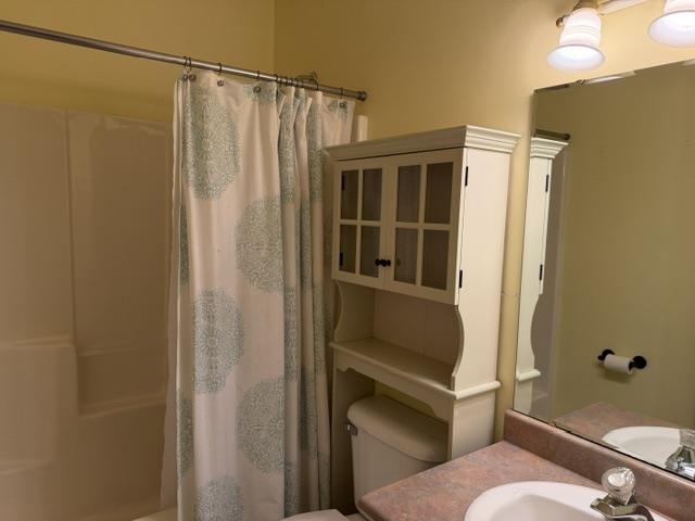 Bathroom featuring a shower/tub combination, wall-mounted vanity lighting, a built-in over-toilet cabinet with glass-front doors, and a vanity with a drop-in sink