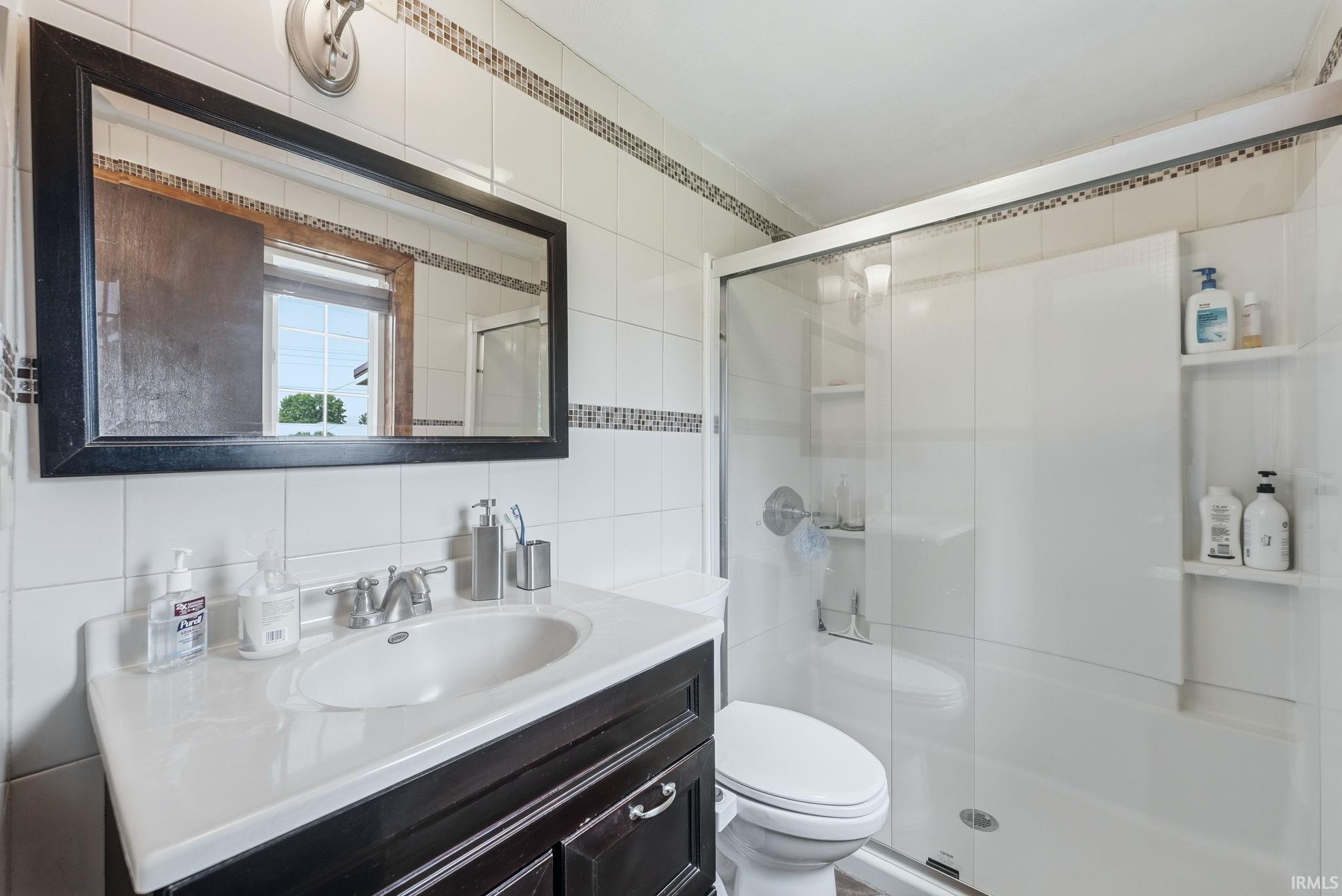 Bathroom featuring a dark wood-finish vanity with a white countertop and integrated sink, a large framed mirror, a tiled shower with a sliding glass door, and recessed shelving within the shower