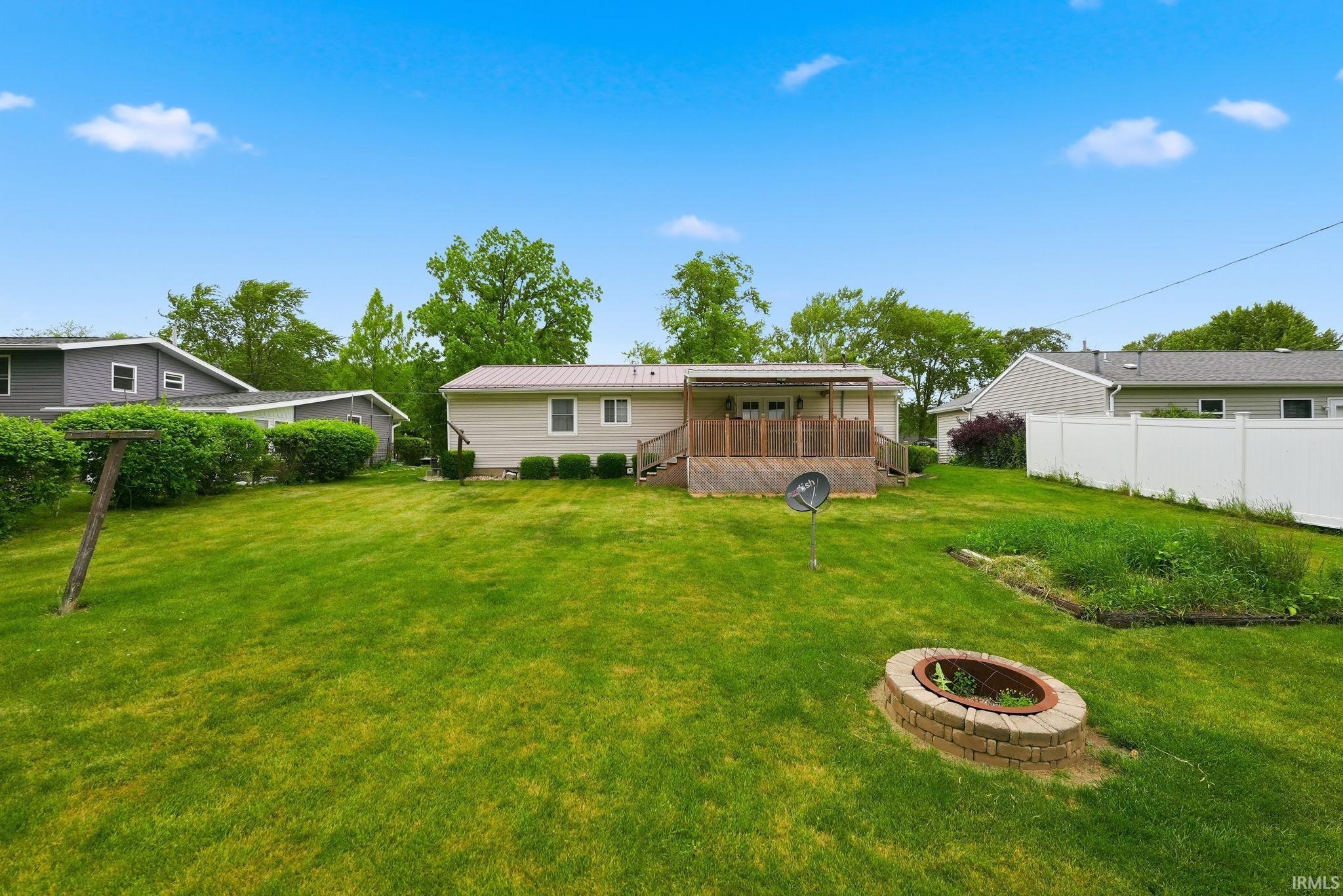 Expansive green lawn featuring a brick-ringed fire pit, a wood deck with a partial covering, and mature perimeter landscaping