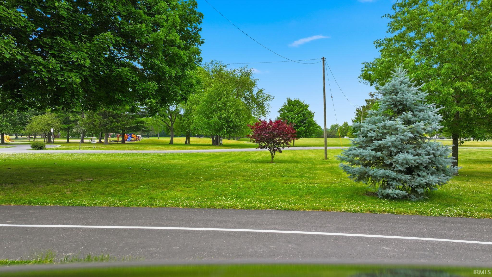 Paved pathway bordering a verdant park space featuring a variety of mature trees and open grassy areas