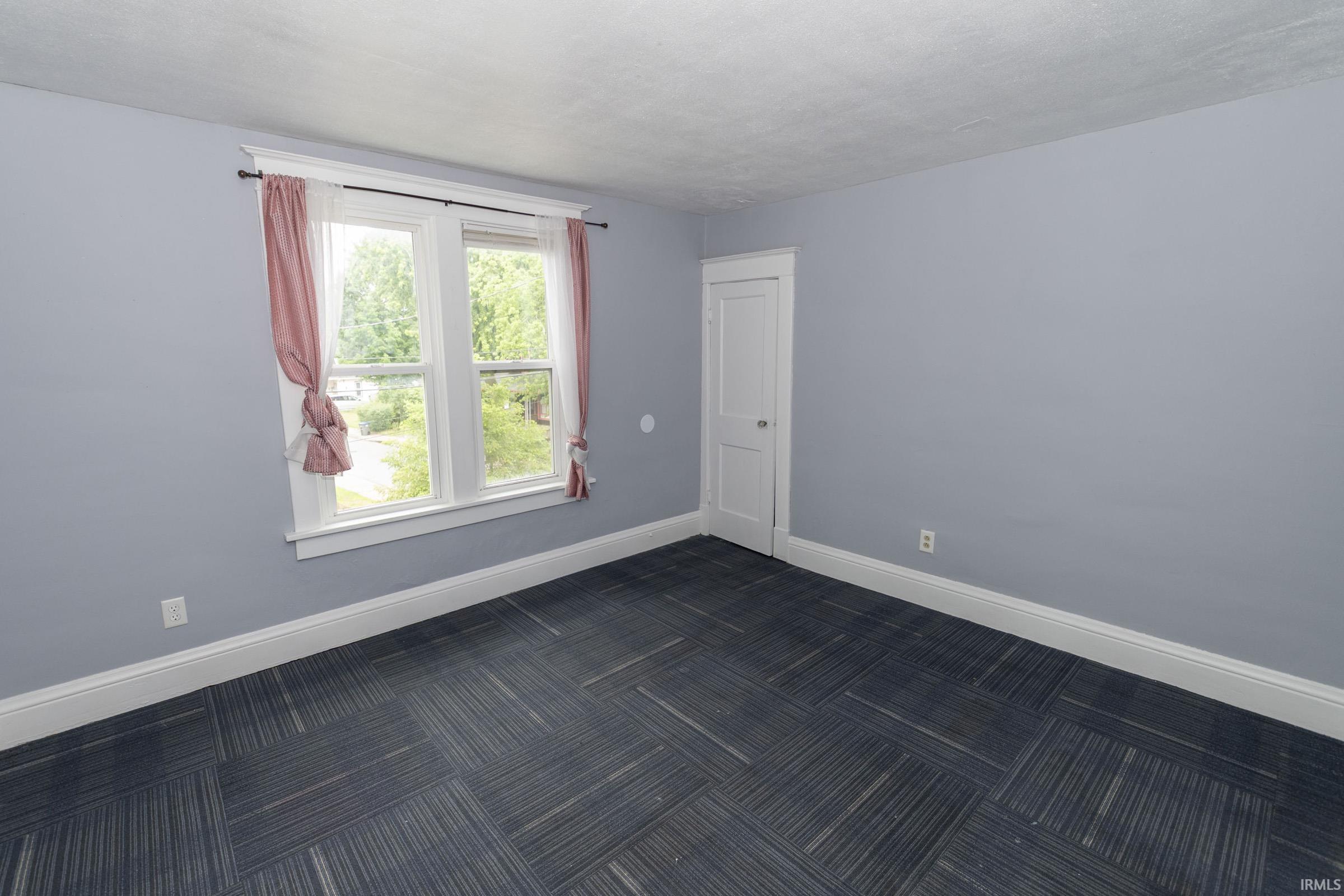 Room featuring a window with white trim, light blue walls, white baseboards, and patterned carpet tile flooring