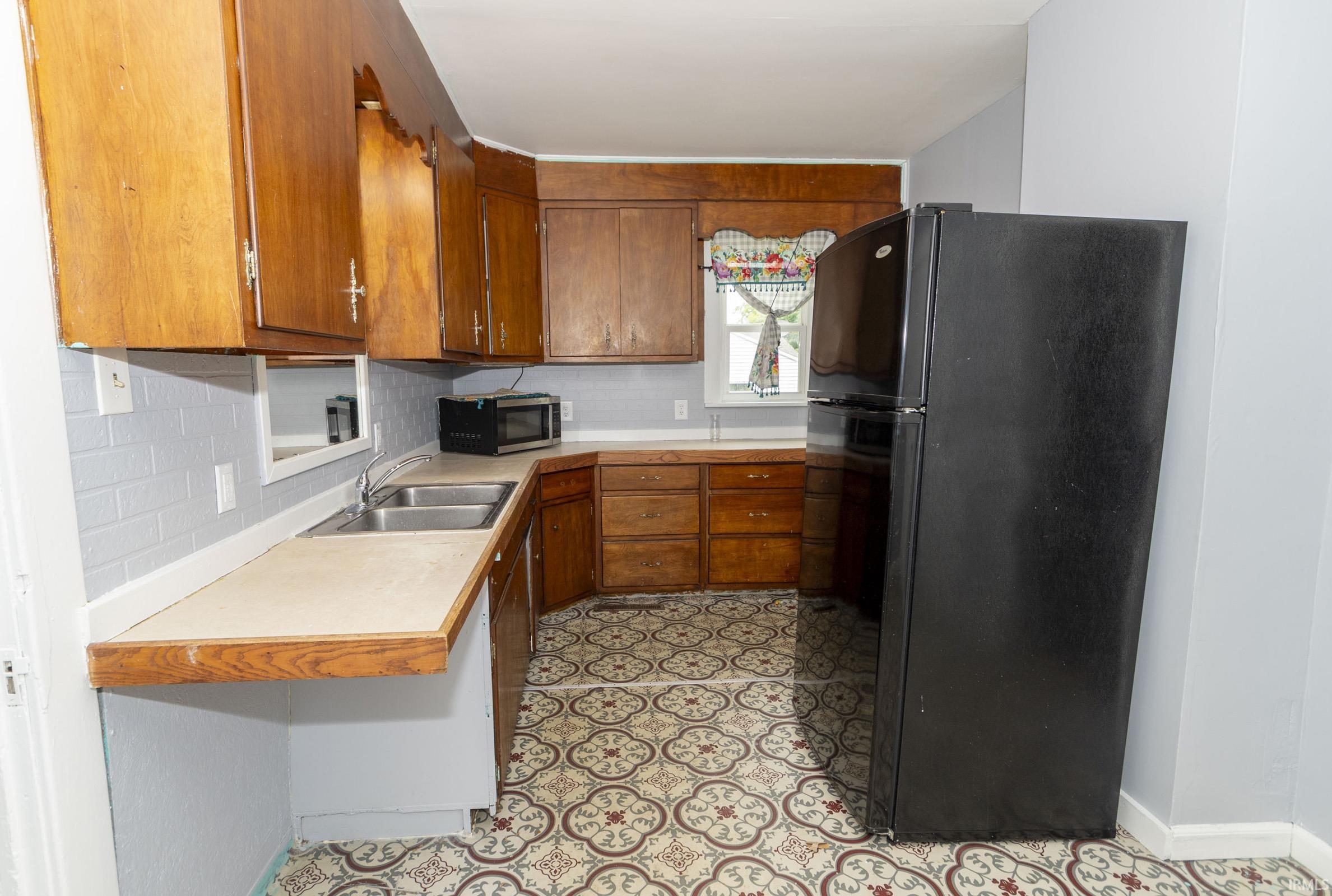 Kitchen featuring patterned floor tile, wood-finish cabinetry, a double basin stainless steel sink, light-toned countertops, and a black refrigerator