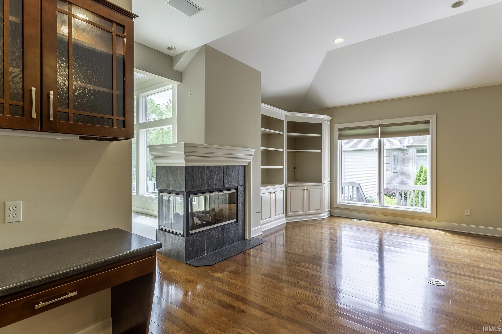 Hardwood flooring throughout this room featuring a corner-oriented three-sided fireplace with dark stone surround