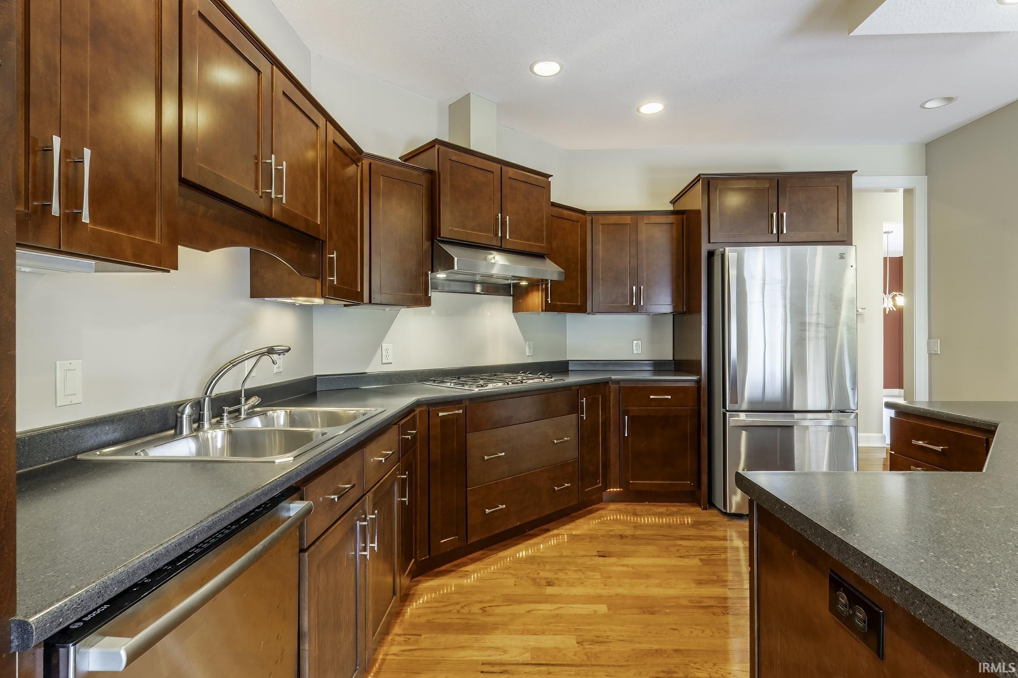Kitchen featuring wood-finish flooring, dark wood cabinetry with silver hardware, stone-look countertops, and stainless steel appliances including a double-basin sink, dishwasher, range with hood, and refrigerator