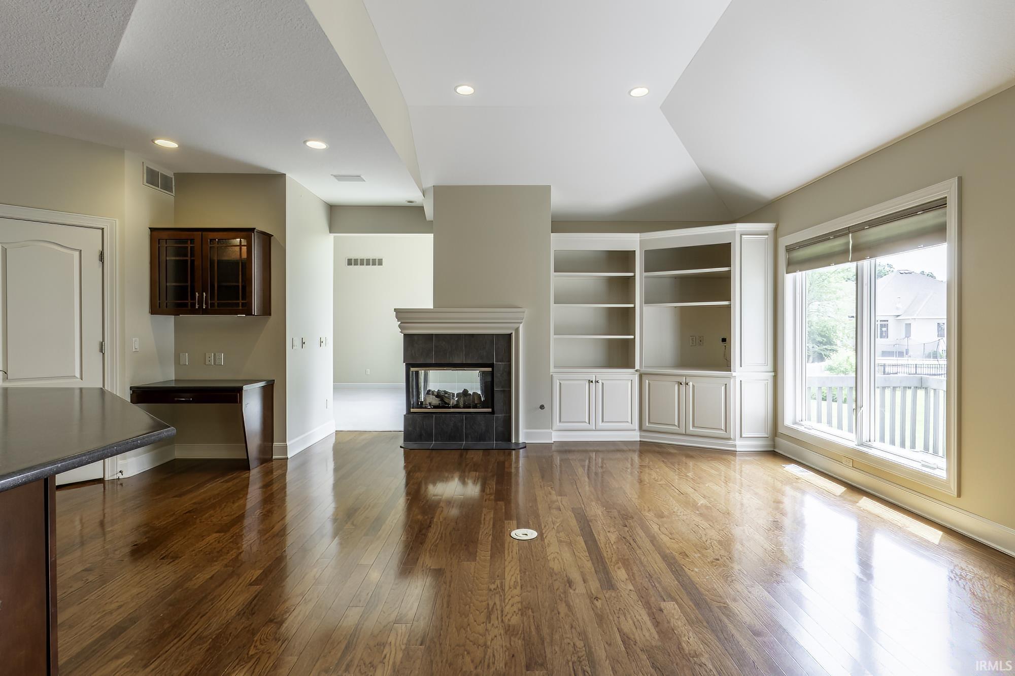 Spacious room featuring wood-finish flooring, a fireplace with tile surround, built-in shelving and cabinetry, a built-in desk with overhead cabinetry, and large windows