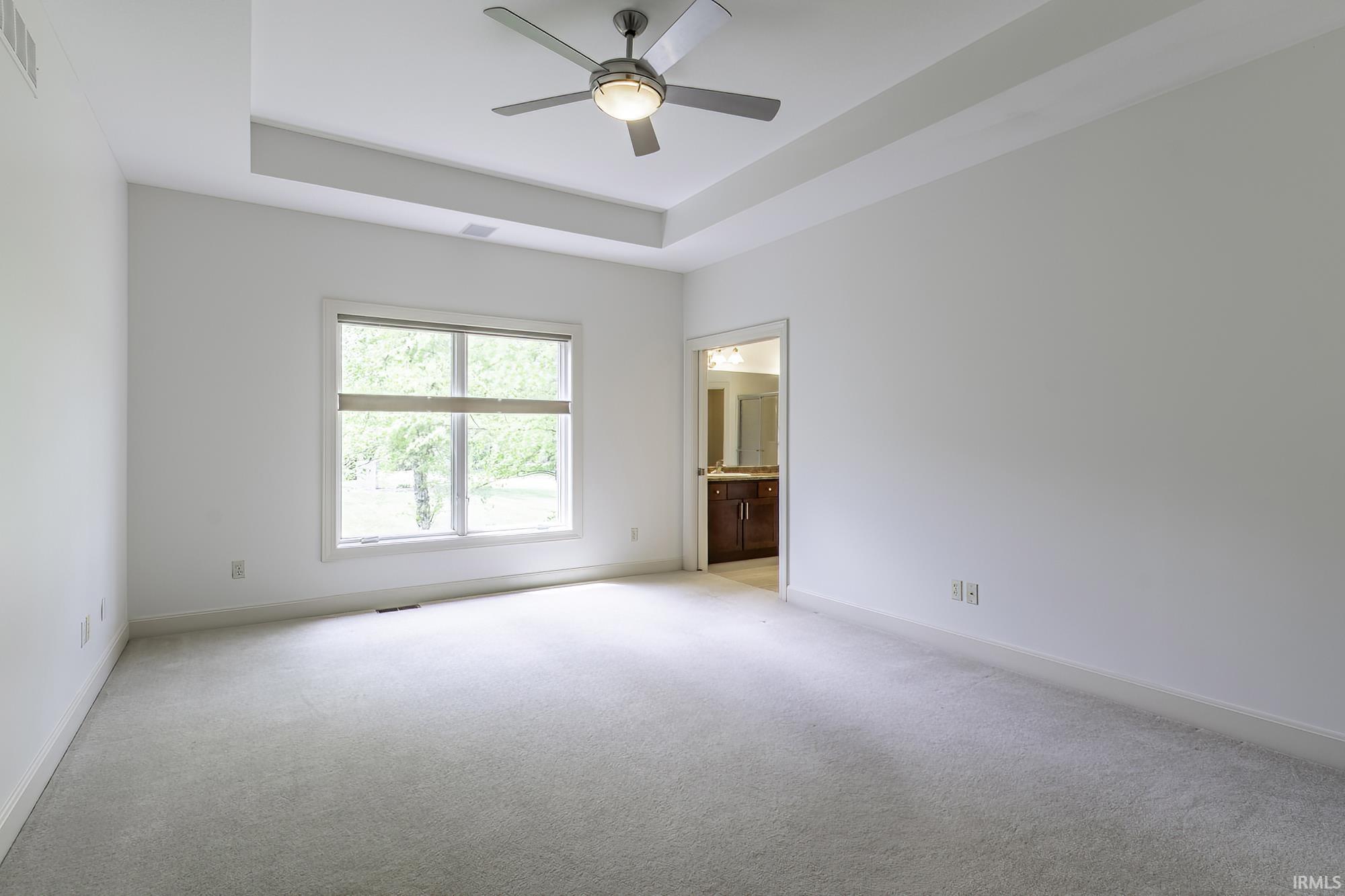 Spacious room featuring a tray ceiling with a ceiling fan, plush carpeting, large window, and an en-suite bathroom with a dark wood vanity
