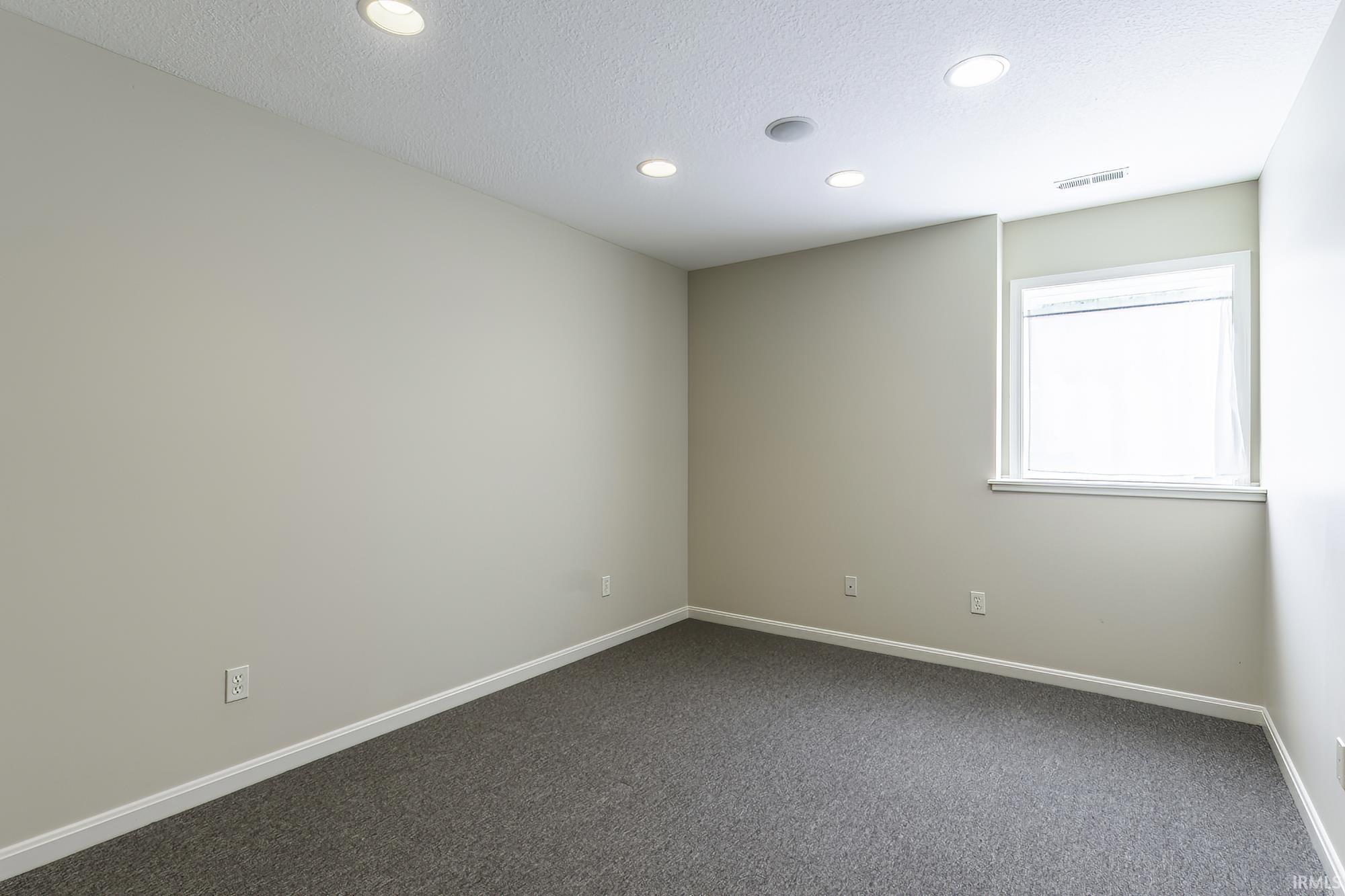Spacious room featuring neutral painted walls, gray carpet flooring, recessed ceiling lighting, and a single window with white trim