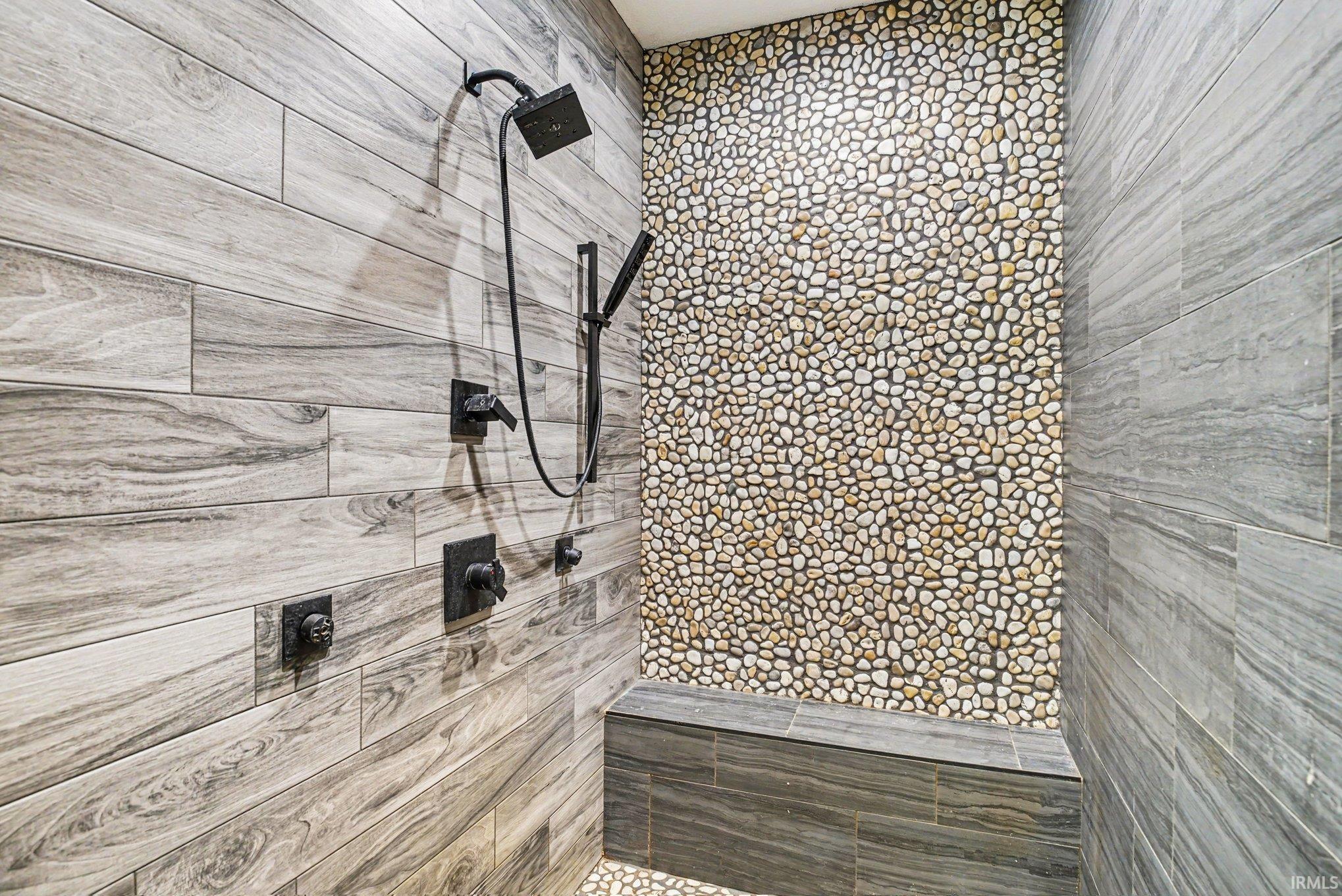 Walk-in shower featuring wood-finish tile walls, pebble accent wall, dark bronze fixtures, a rainfall showerhead, and a built-in bench