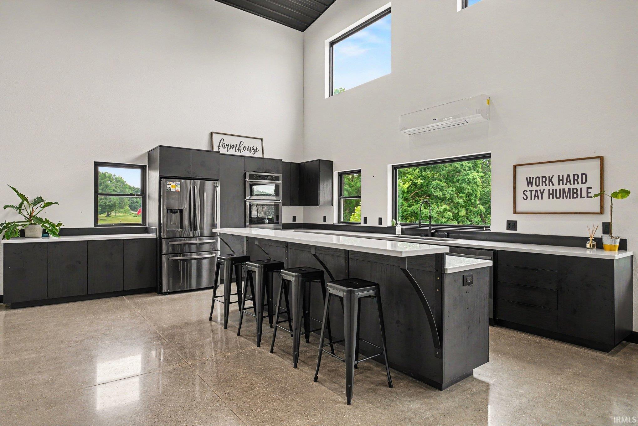Modern kitchen featuring polished concrete flooring, extensive dark cabinetry with light countertops, stainless steel appliances, and high ceilings