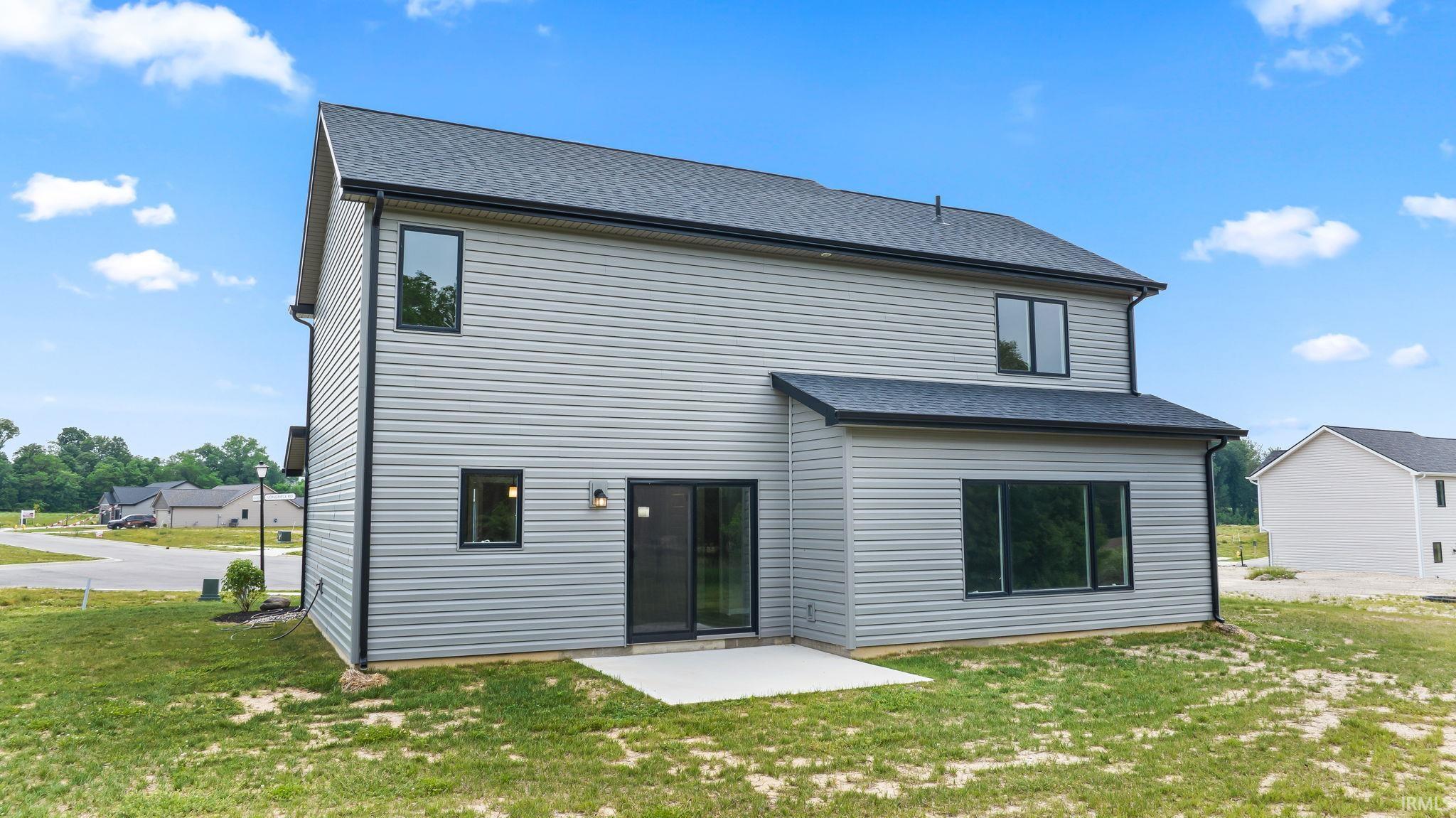 Rear exterior featuring light gray siding, dark-framed windows, a black shingled roof, a ground-level concrete patio, and a sliding glass door