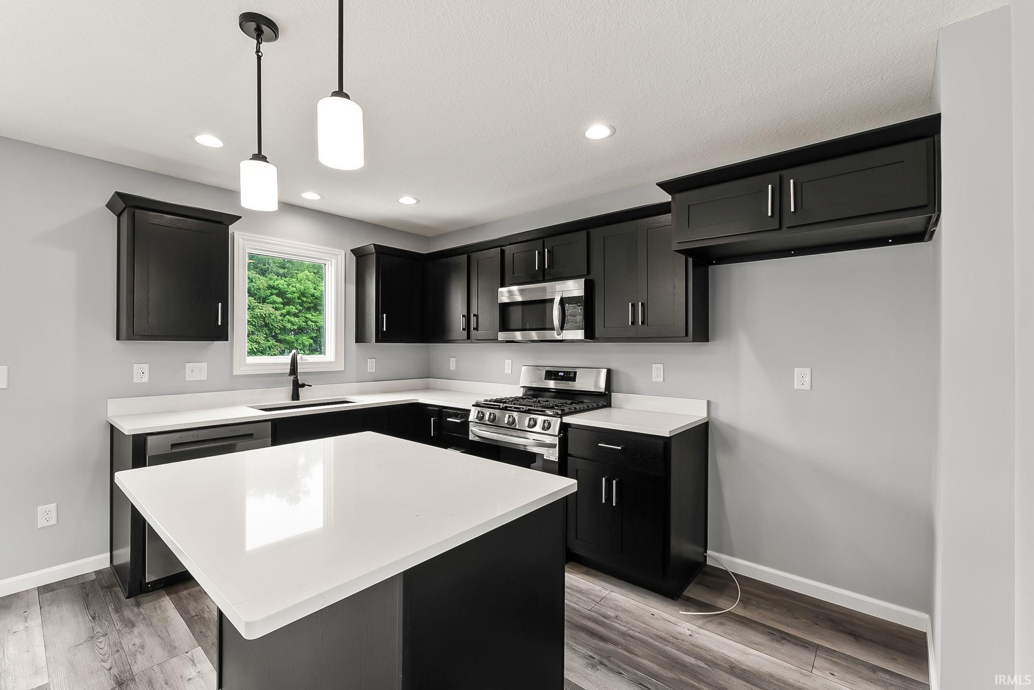 Modern kitchen featuring a central island with a white countertop, dark cabinetry, stainless steel appliances, wood-finish flooring, and recessed lighting