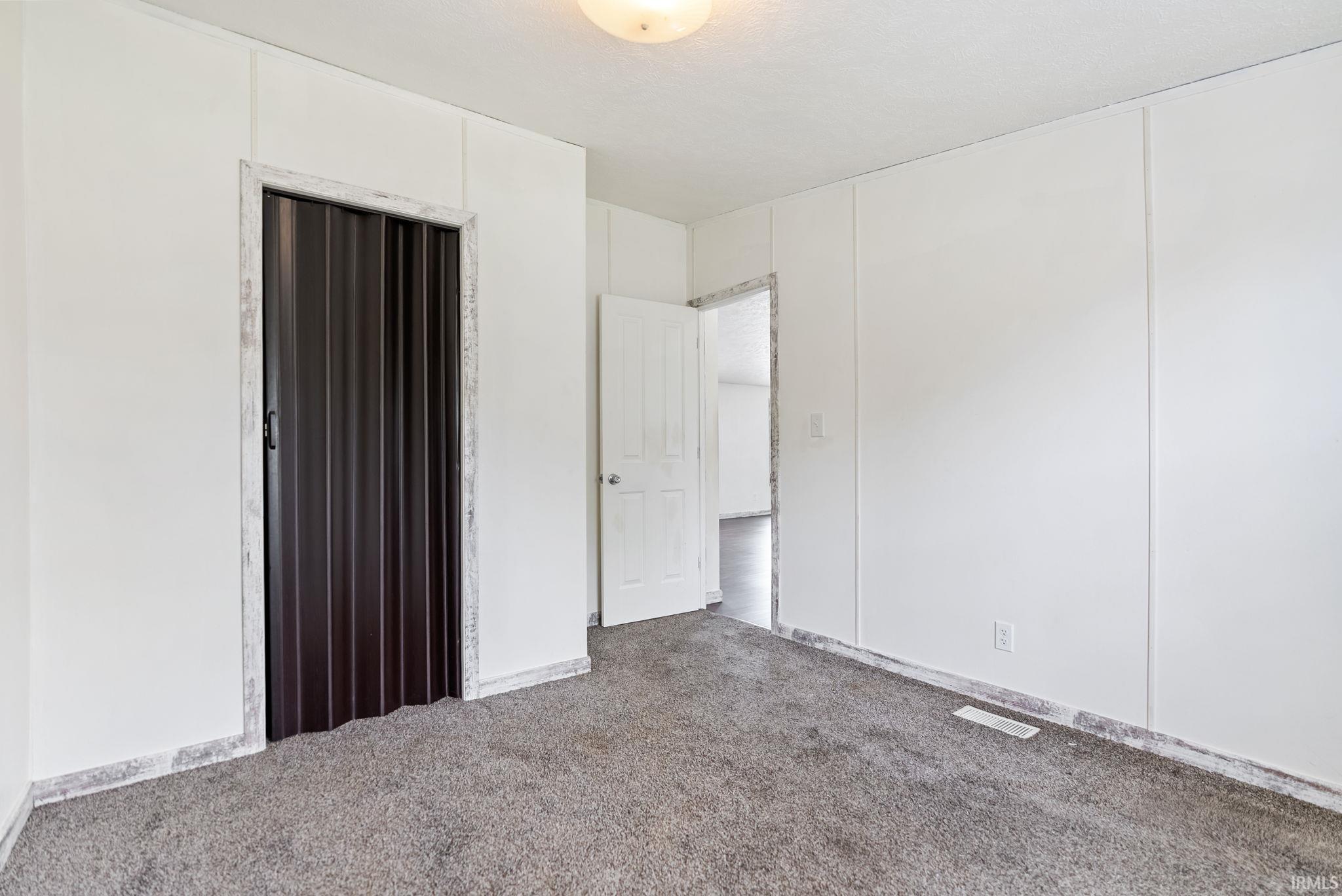Interior room featuring white panel walls, grey carpet flooring, a dark brown accordion door, a white panel door, and ceiling-mounted lighting