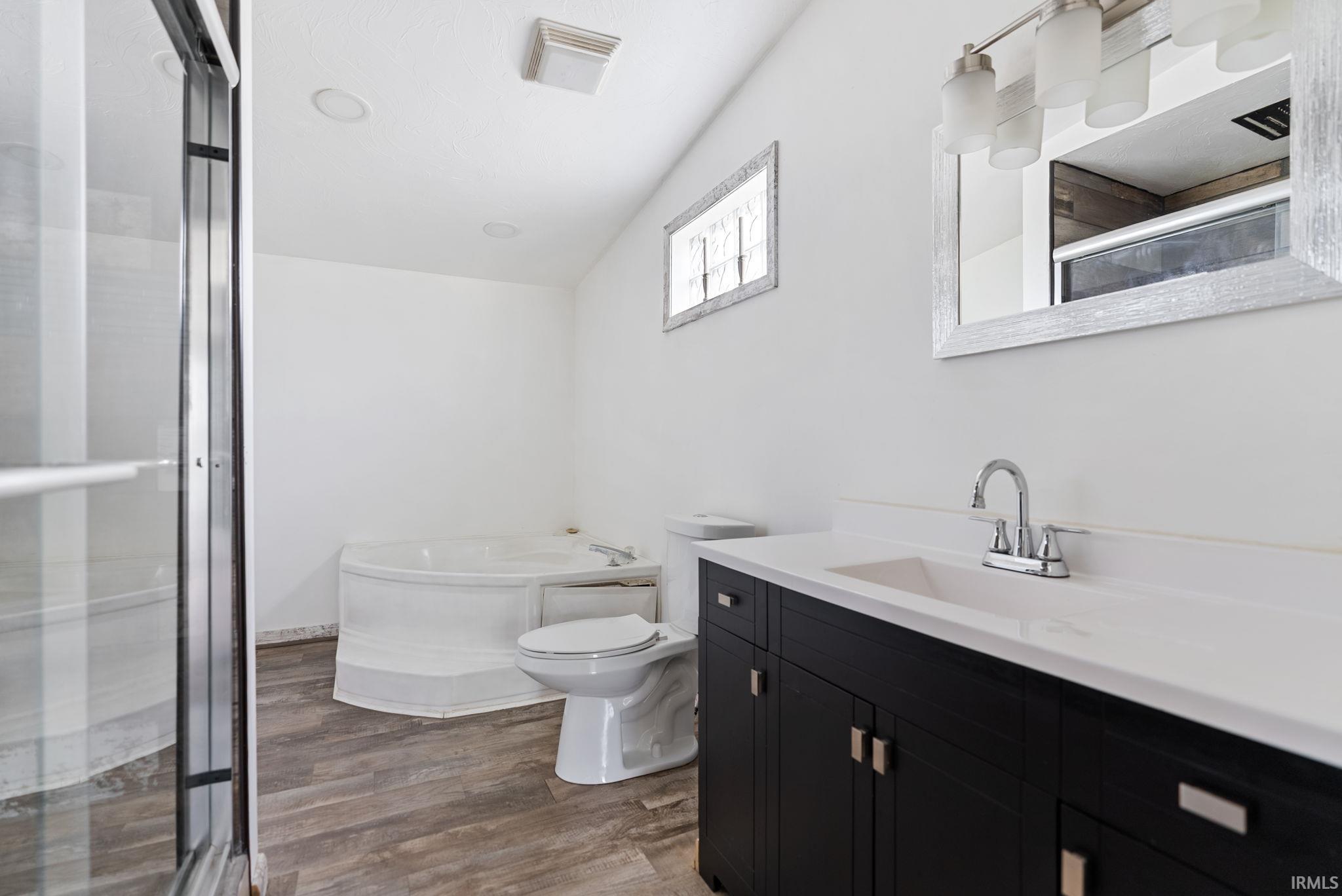 Bathroom featuring a white jetted tub, a dark wood-finish vanity with a white countertop and integrated sink, a large framed mirror, and wood-finish flooring