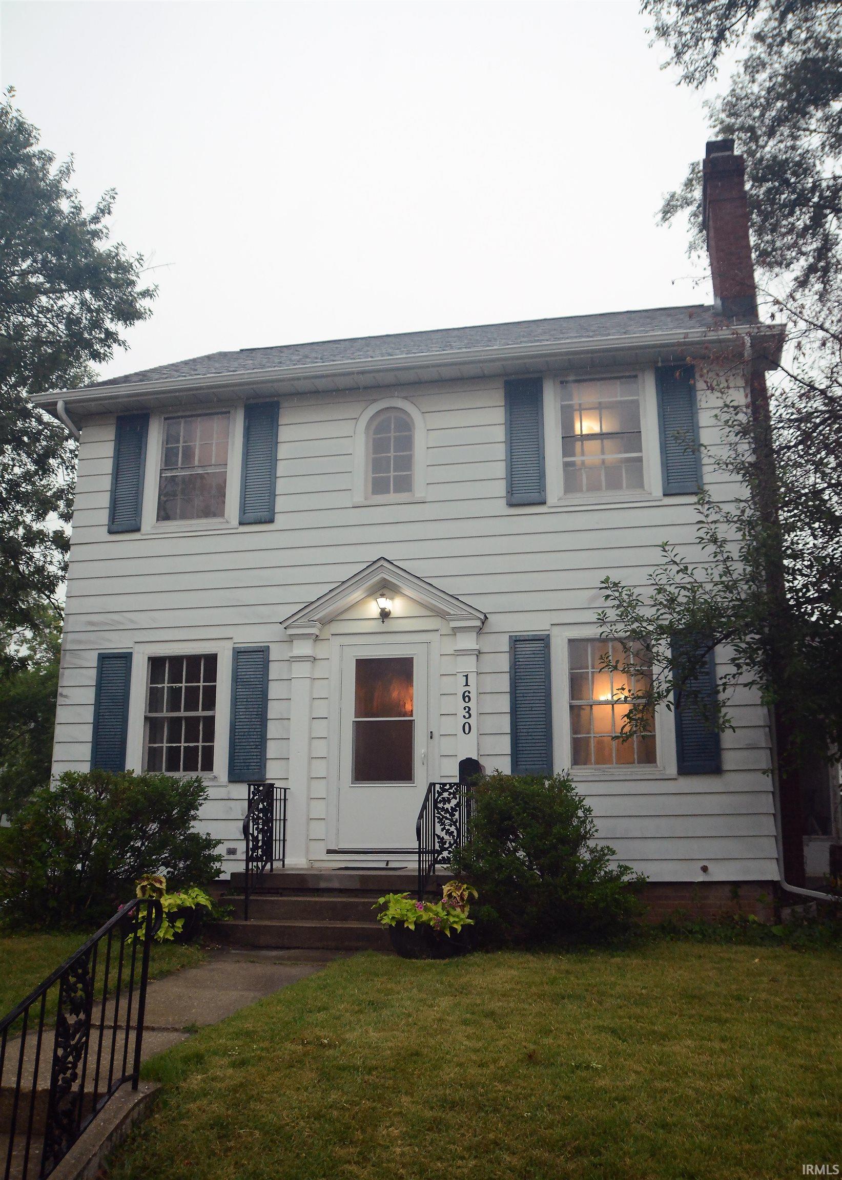 White siding exterior featuring a prominent brick chimney, classic blue shutters, and a decorative arched window