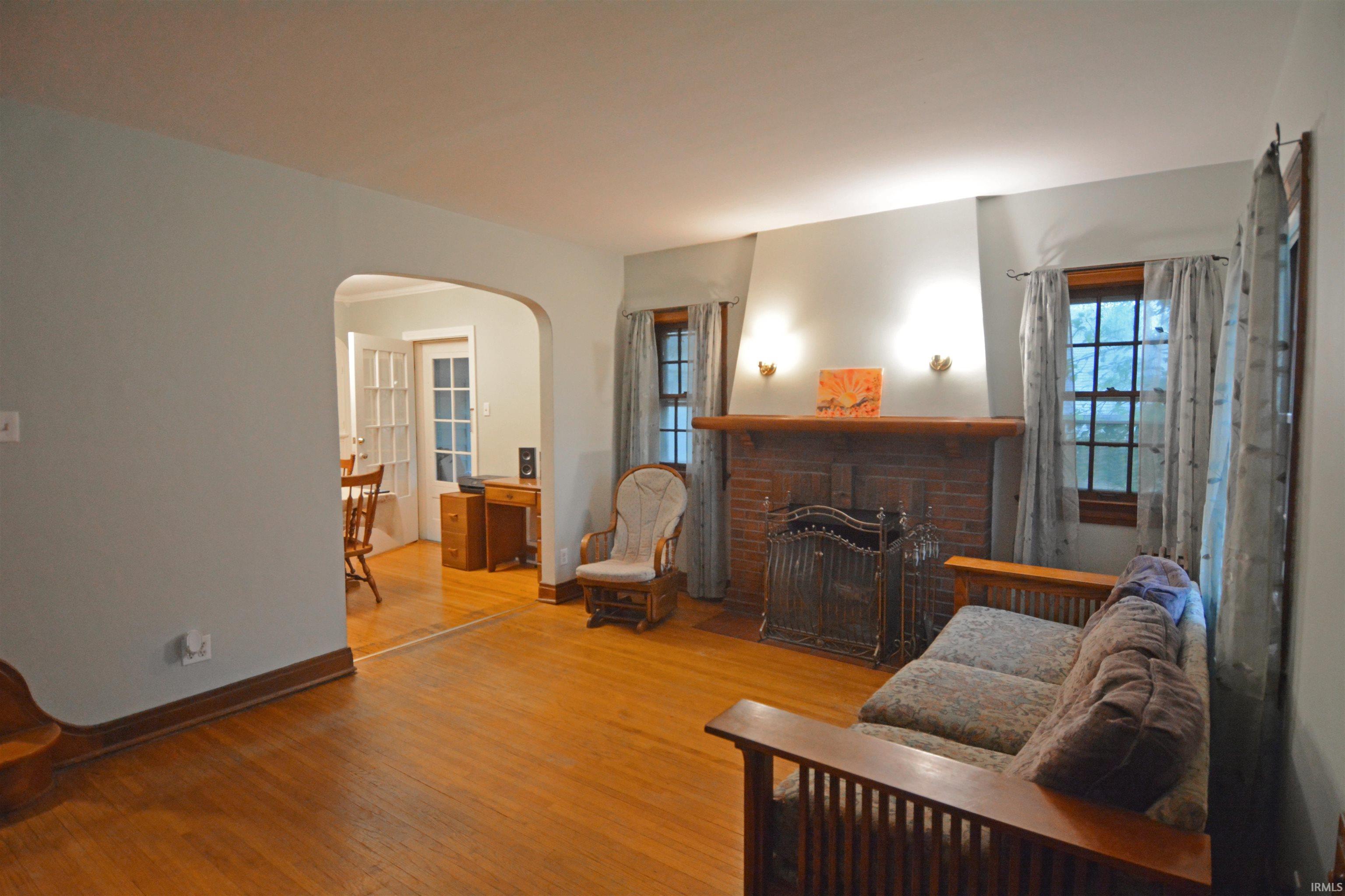 Living area featuring hardwood flooring, a brick fireplace with a wood mantle, twin sconce lighting, and an arched doorway opening to an adjacent room