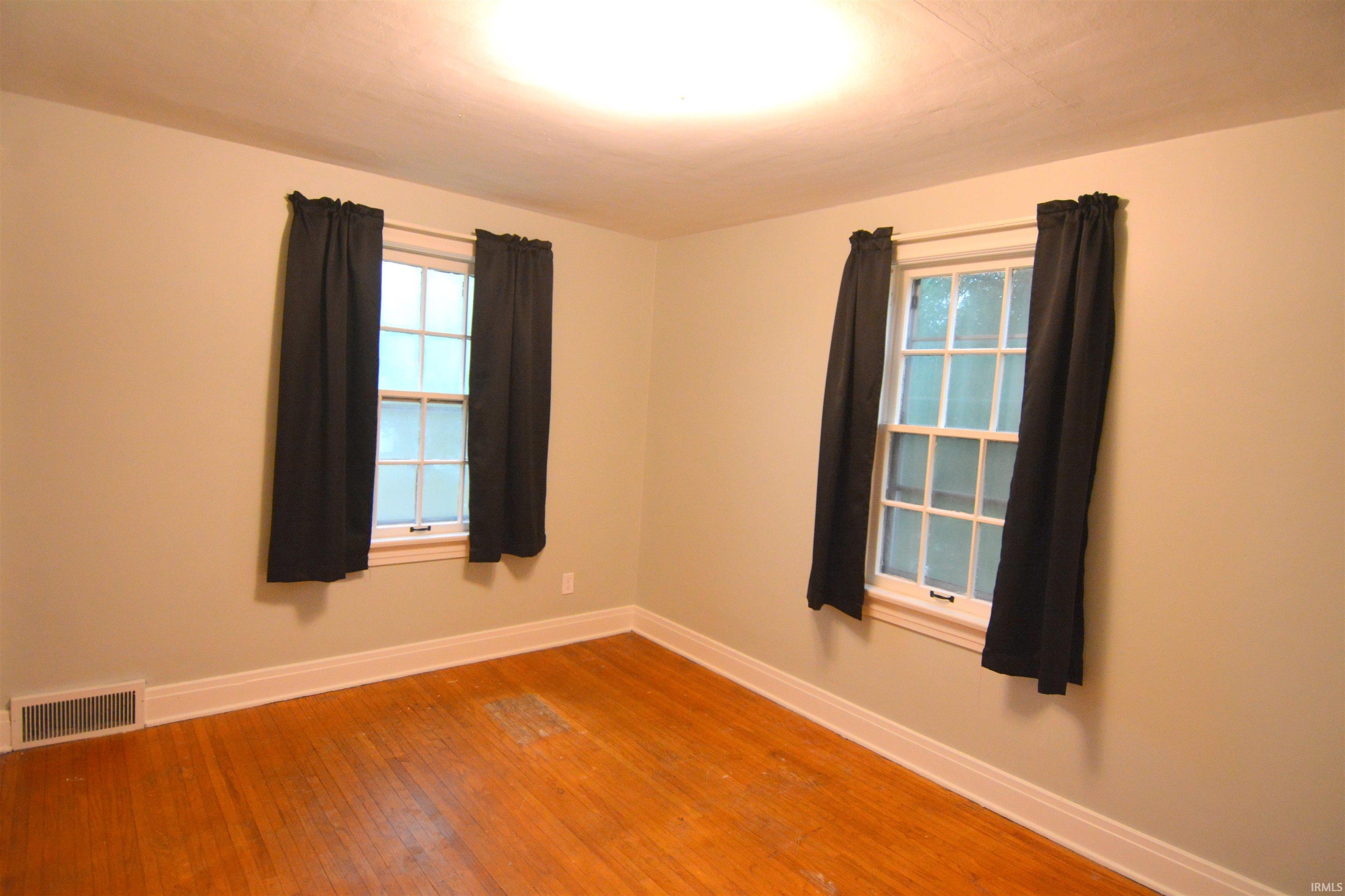 Hardwood flooring extends throughout this room, featuring two windows with white frames and dark curtains
