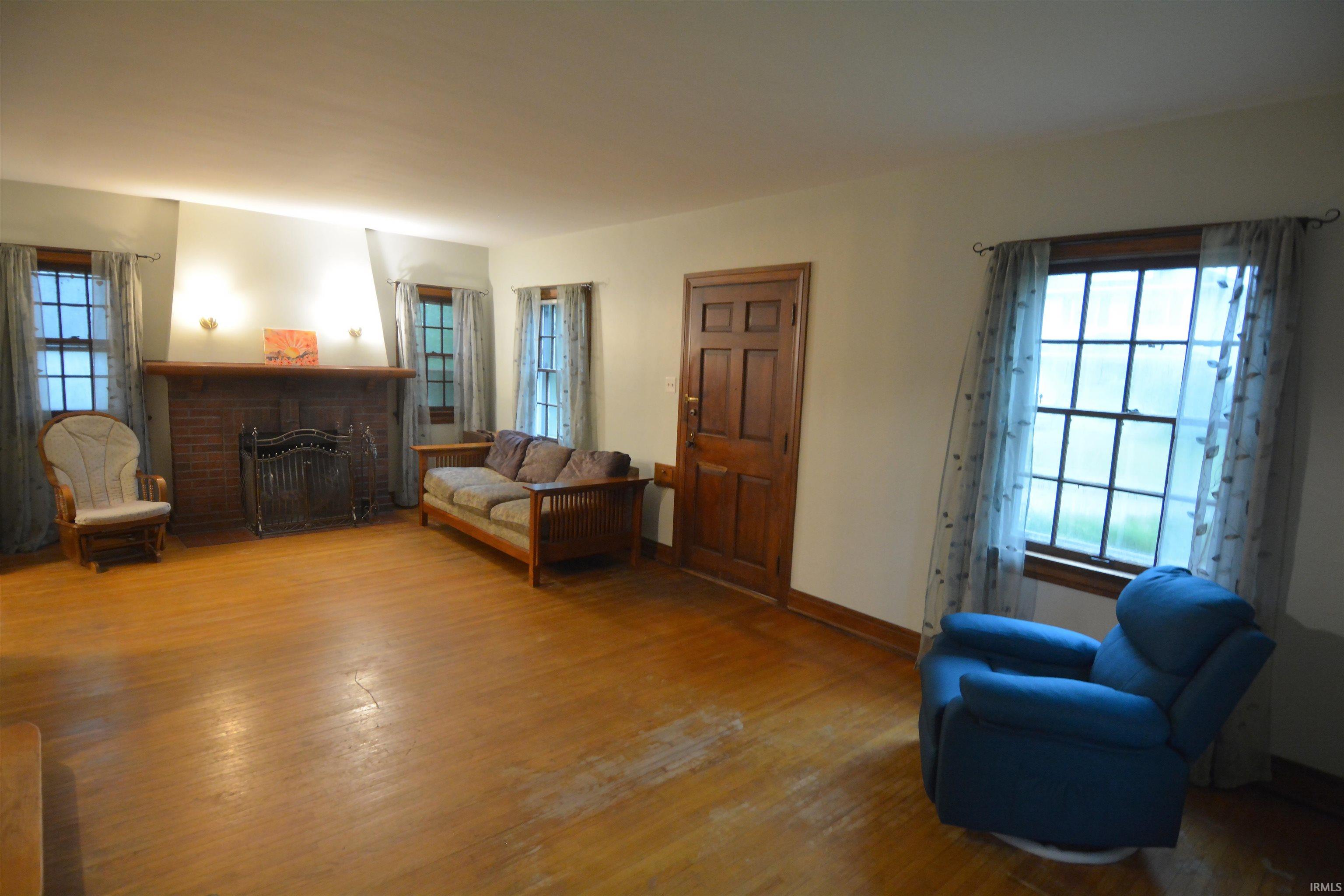 Spacious living area featuring wood-finish flooring, a brick fireplace with a wood mantel, multiple pane windows, a substantial paneled door, and wall sconces