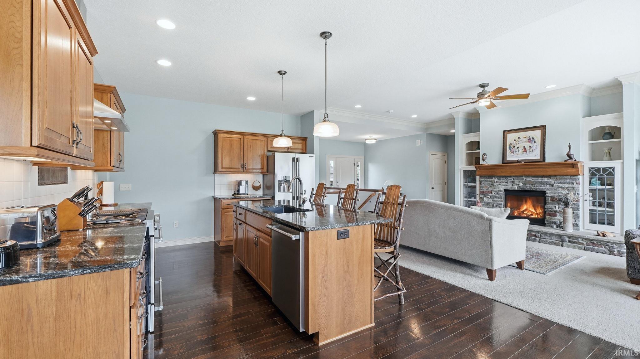 Open concept living area featuring dark wood-finish flooring in the kitchen, a stone fireplace with a wood mantle, custom built-in shelving, recessed lighting, and a ceiling fan