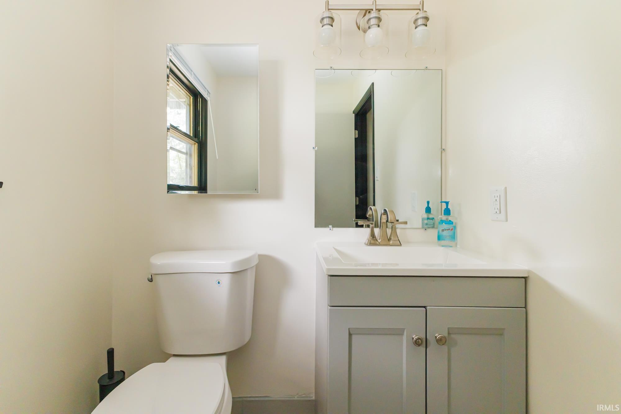 Bathroom featuring a white toilet, gray vanity with two cabinet doors, an integrated white sink, a brushed nickel faucet, and overhead three-bulb vanity lighting