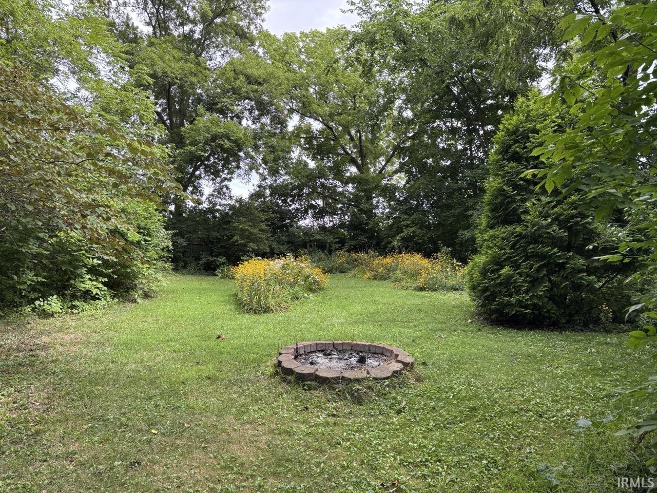 Expansive backyard featuring a central brick-ringed fire pit, manicured lawn, and mature perimeter trees providing natural screening