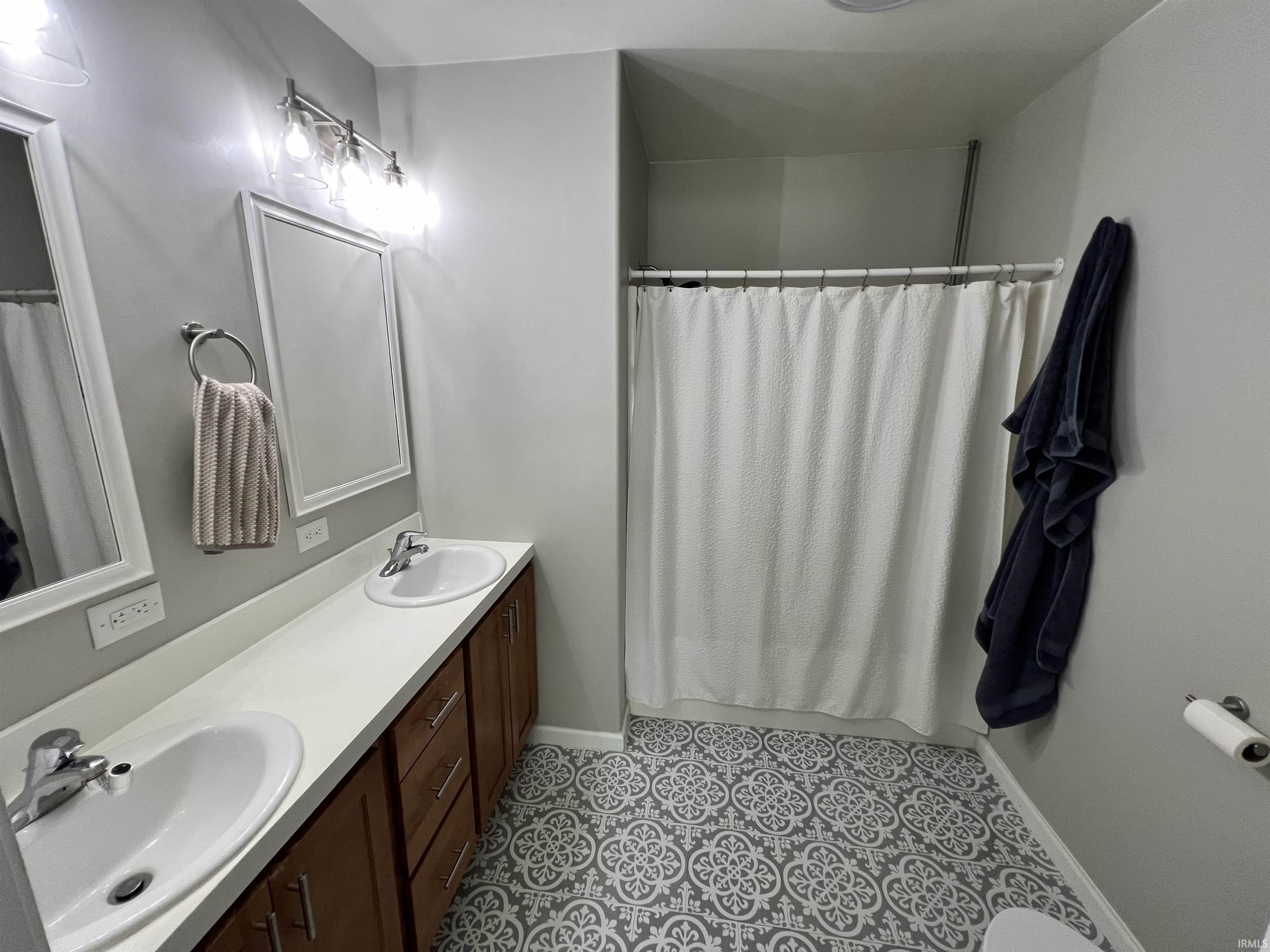 Bathroom with dual vanity, white countertops, wood-finish cabinetry, and patterned floor tiling