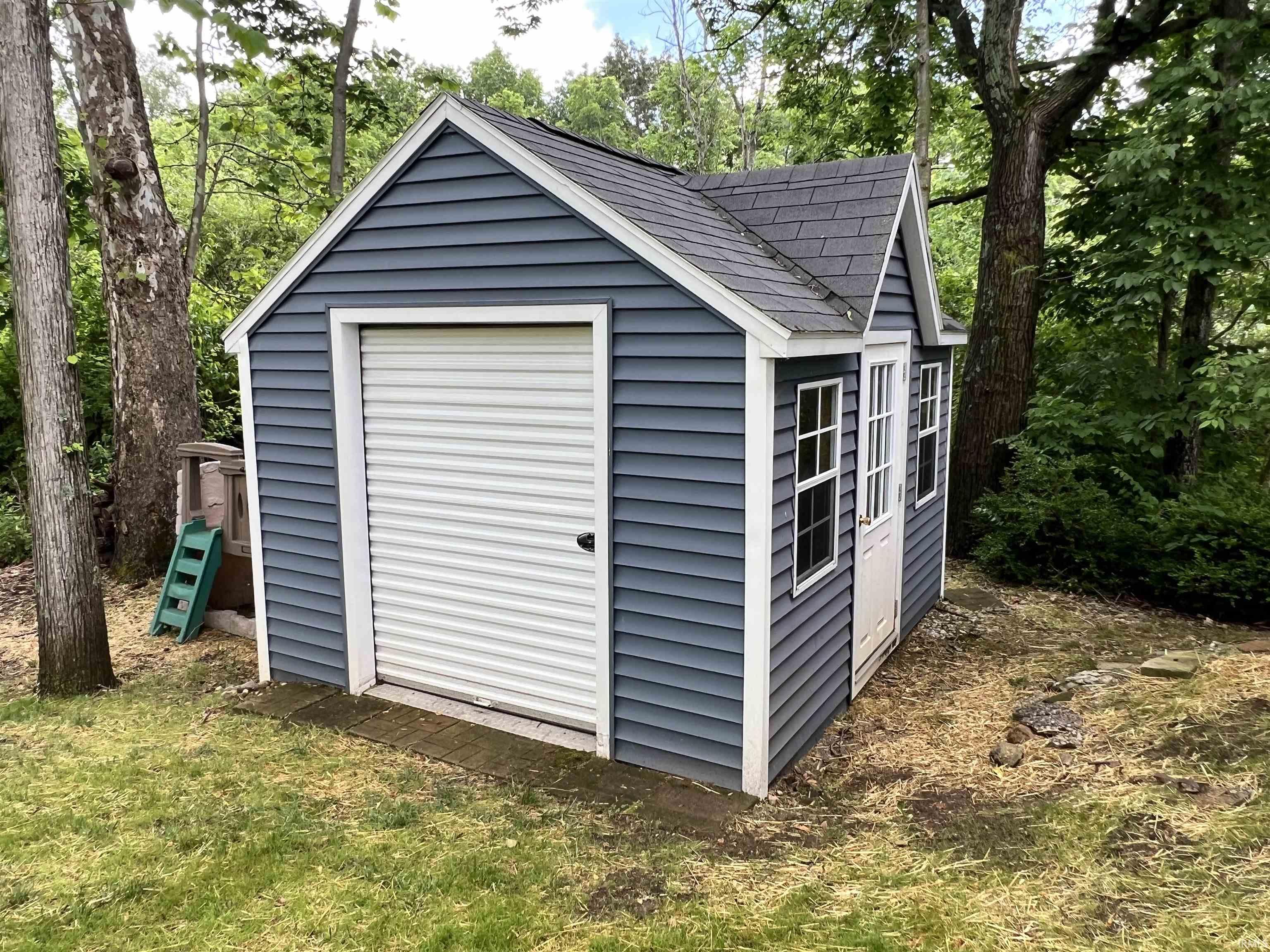 Freestanding shed featuring blue horizontal siding, white trim, a black shingled roof, a roll-up garage door, and a white side entry door with two windows