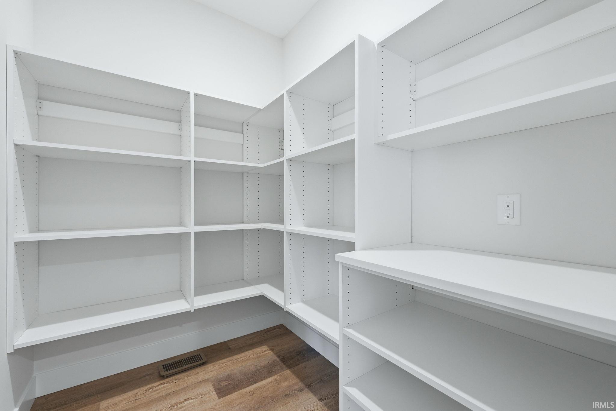 Walk-in pantry featuring extensive white built-in shelving, wood-finish flooring, and a wall outlet for convenience