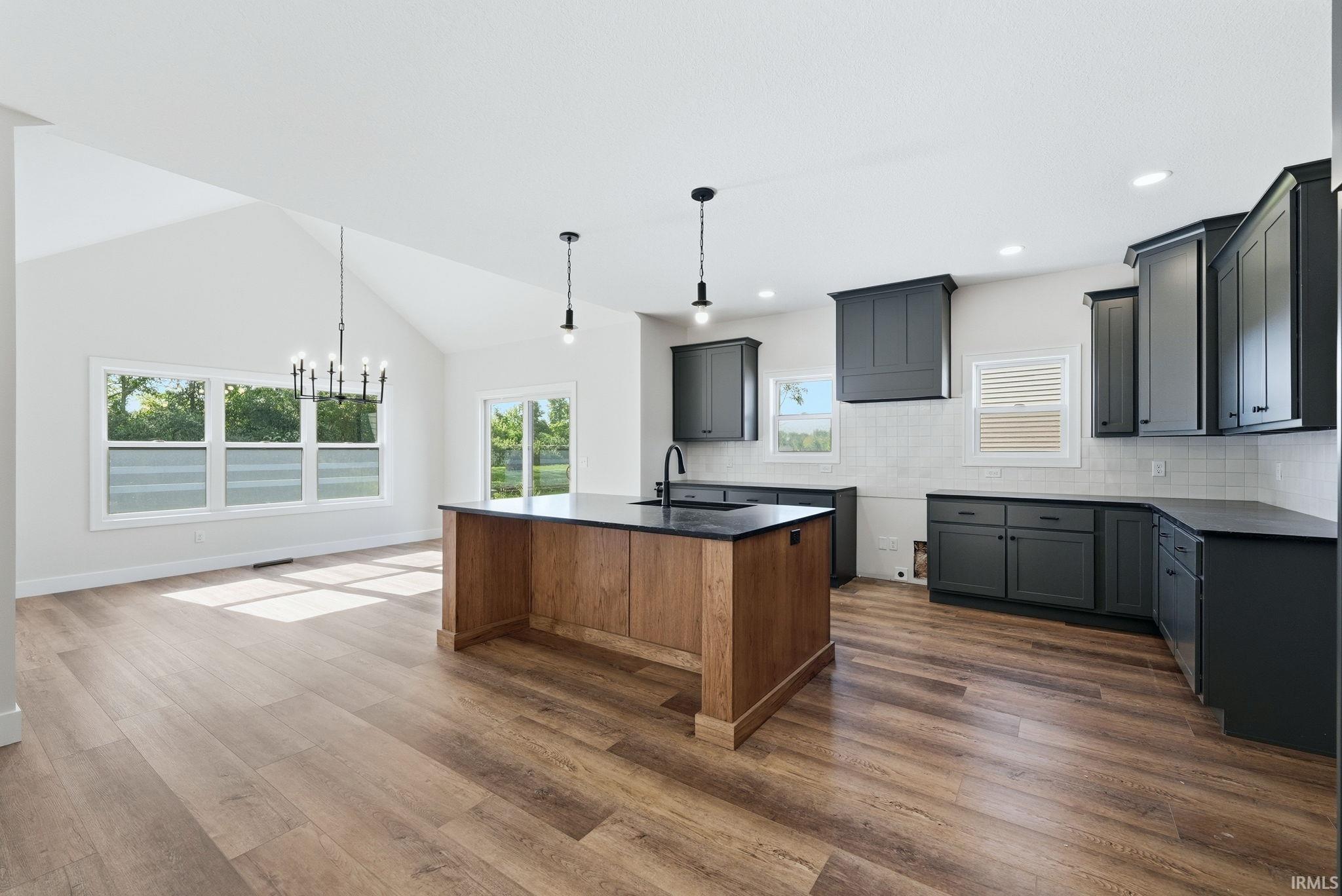 Open-concept kitchen and dining area featuring wood-finish flooring, a vaulted ceiling, a kitchen island with dark countertops, dark shaker-style cabinetry, and a white subway tile backsplash