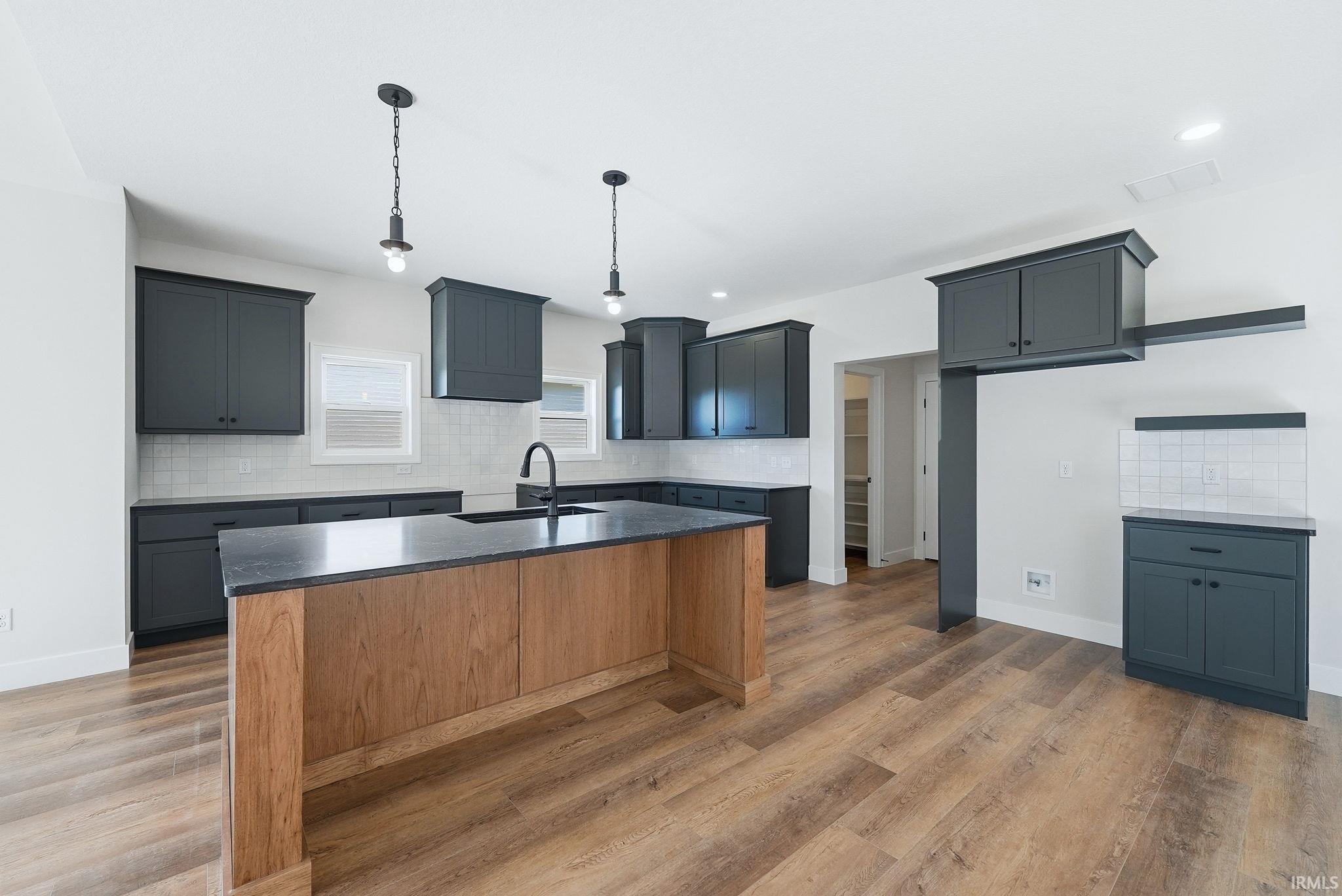 Modern kitchen featuring wood-finish flooring, dark gray cabinetry, a central island with wood-finish paneling and a dark countertop, white subway tile backsplash, and matte black fixtures