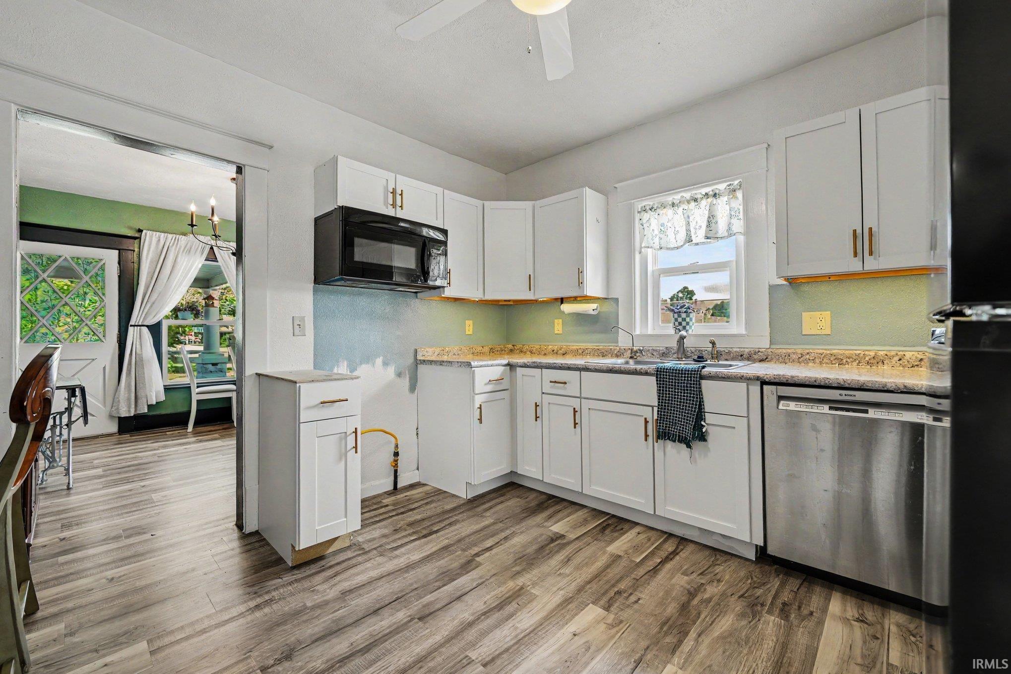 Kitchen featuring wood-finish flooring, white cabinetry with gold-finish hardware, a stainless steel dishwasher, an over-range microwave, and a window above the sink