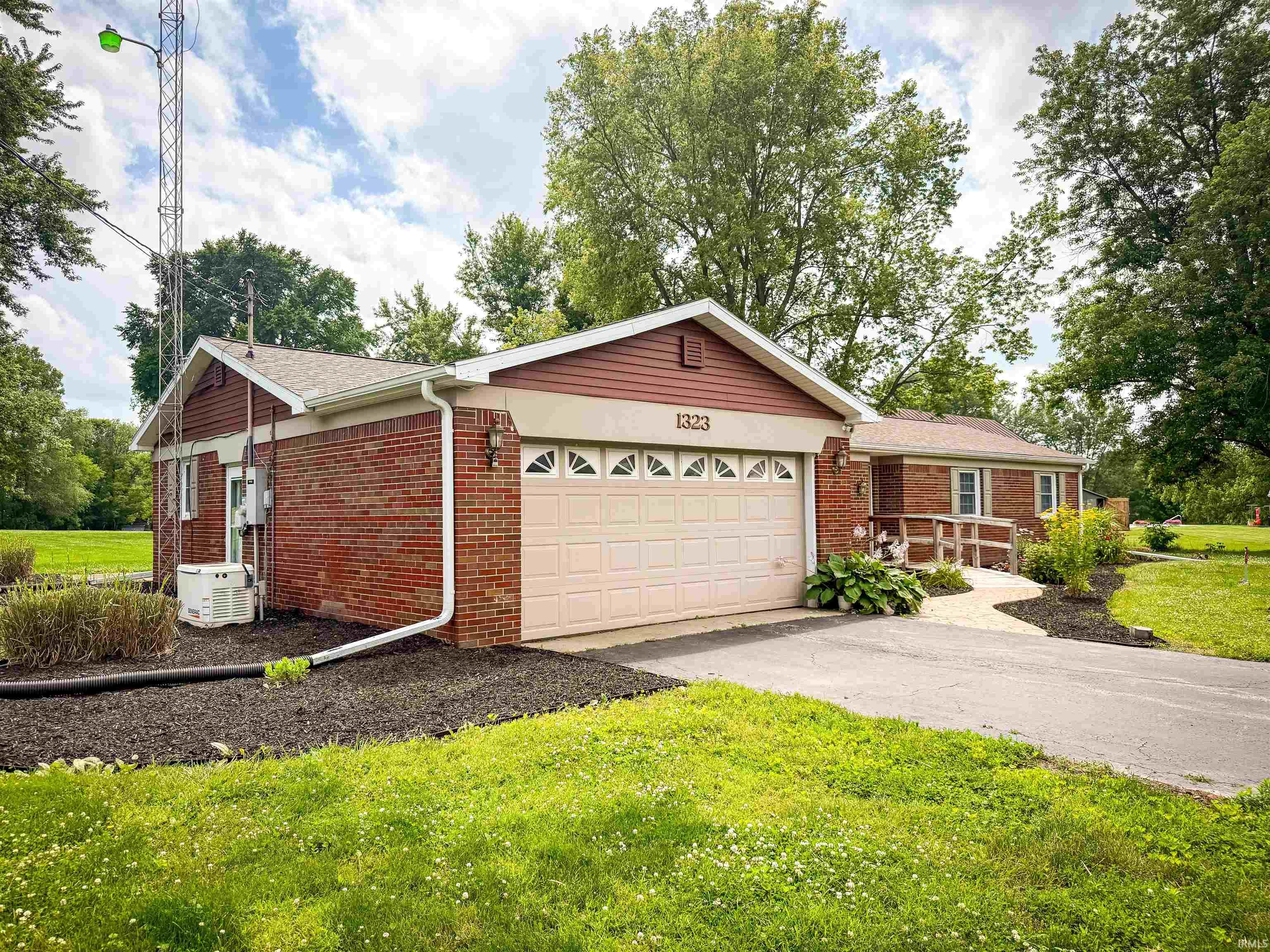 Brick exterior residence featuring a two-car garage with a paneled door, a paved driveway, a landscaped front yard with dark mulch, and a front entry ramp