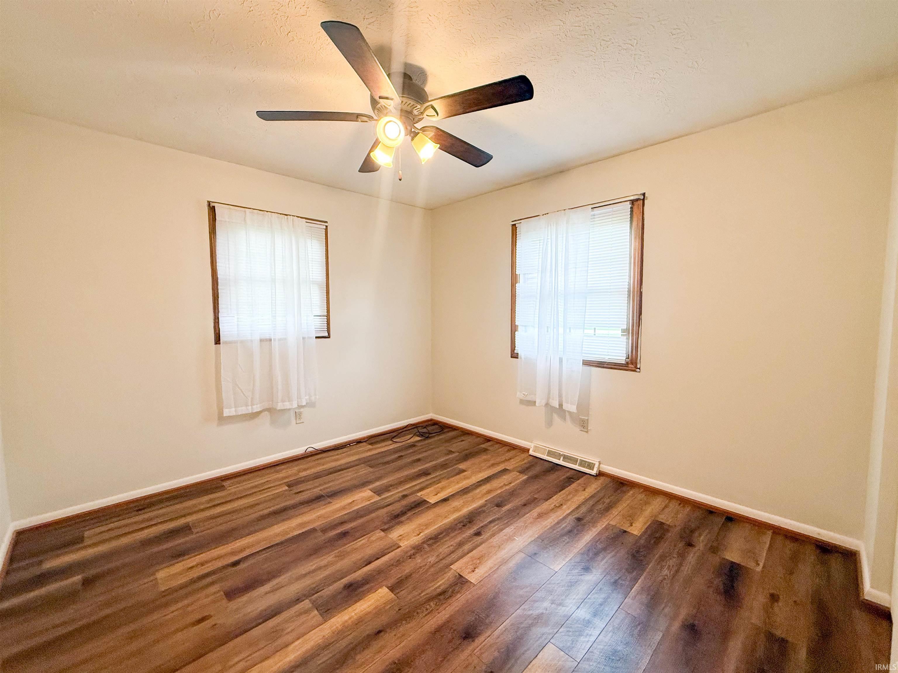 Room featuring wood-finish flooring, two windows with blinds and sheer curtains, a ceiling fan with integrated lighting, and neutral wall tones